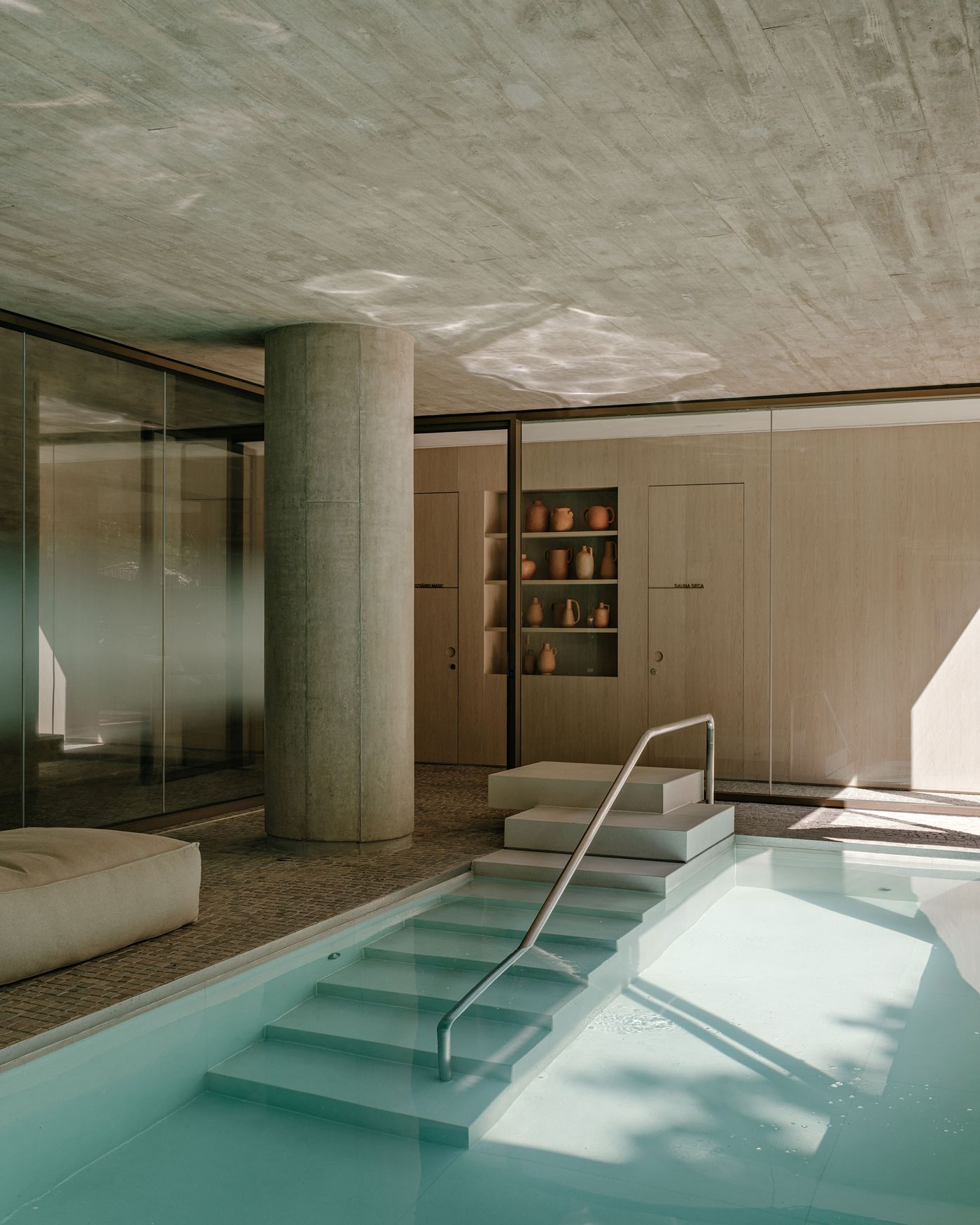 The serene indoor pool area, showcasing a dramatic play of sunlight filtering through the space, creating captivating patterns on the raw concrete ceiling and water. The minimalist concrete columns and warm wooden cabinetry with curated ceramic vessels offer a spa-like, Zen-inspired atmosphere. The serene indoor pool area, showcasing a dramatic play of sunlight filtering through the space, creating captivating patterns on the raw concrete ceiling and water. The minimalist concrete columns and warm wooden cabinetry with curated ceramic vessels offer a spa-like, Zen-inspired atmosphere.