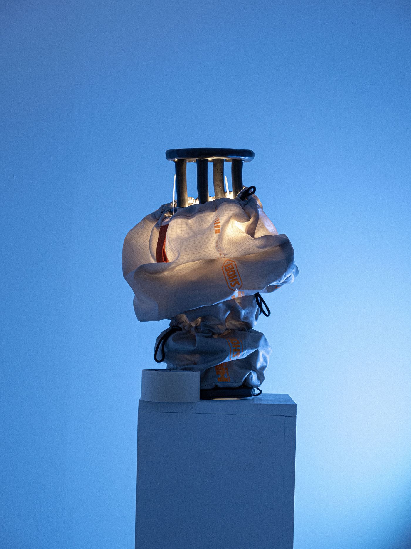 A shiny metal object in a white bag, illuminated against a blue backdrop, rests on a pedestal with tape beside it. A shiny metal object in a white bag, illuminated against a blue backdrop, rests on a pedestal with tape beside it.