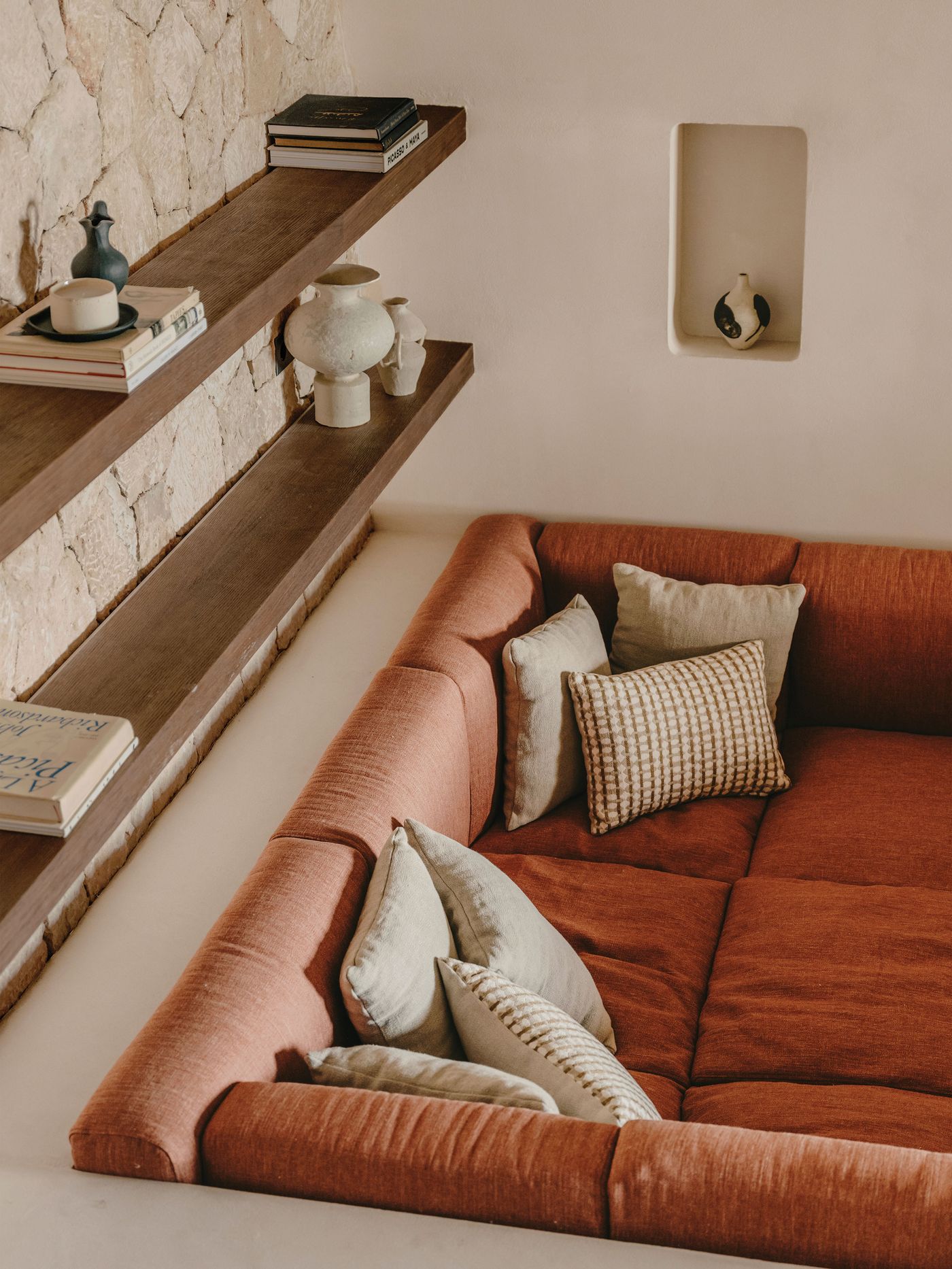 A cozy rust-colored sofa adorned with various patterned pillows, beside wooden shelves displaying pottery and books against a stone wall. A cozy rust-colored sofa adorned with various patterned pillows, beside wooden shelves displaying pottery and books against a stone wall.