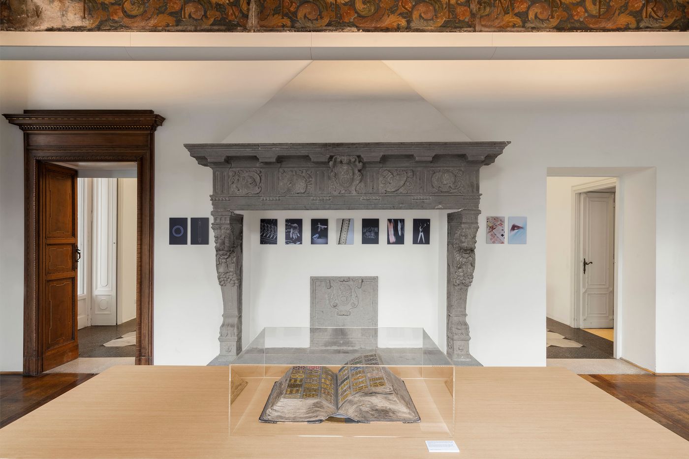 An ornate, carved stone fireplace serves as the focal point of this room, framed by traditional wooden doors. Above the mantelpiece, a series of small framed artworks are displayed. In front of the fireplace, an open, ancient book rests within a modern glass display case on a simple wooden table. The image blends historical architectural elements with contemporary exhibition design, creating a sense of refined presentation.