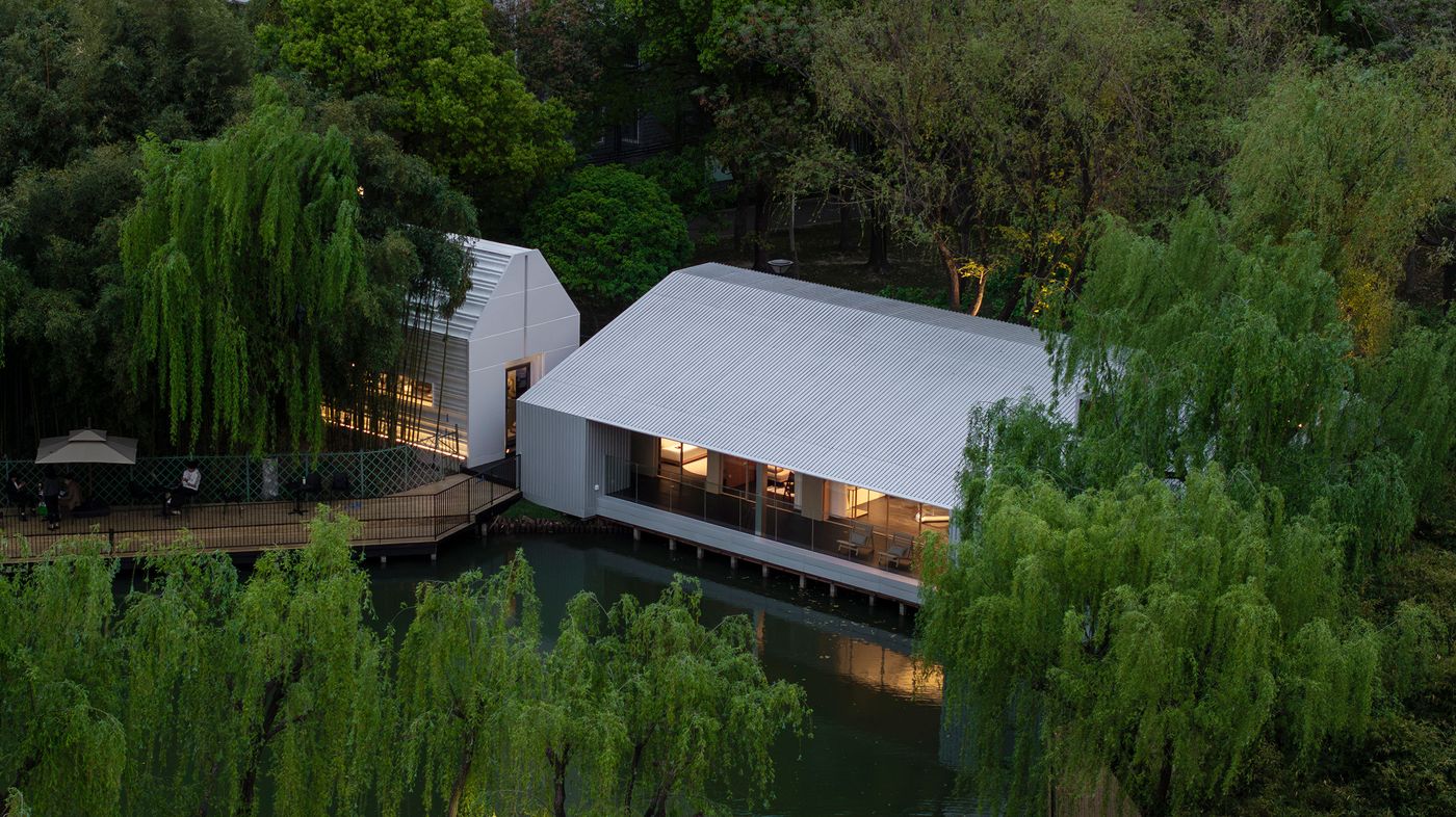 An aerial view at dusk reveals two distinct, gabled forms, their white corrugated metal surfaces subtly reflecting the evening light. Warm glows from the interiors invite contemplation. Surrounded by lush, dark green foliage and set against the tranquil lake, the composition highlights the architectural dialogue between modern forms and natural serenity, creating a harmonious biophilic retreat. An aerial view at dusk reveals two distinct, gabled forms, their white corrugated metal surfaces subtly reflecting the evening light. Warm glows from the interiors invite contemplation. Surrounded by lush, dark green foliage and set against the tranquil lake, the composition highlights the architectural dialogue between modern forms and natural serenity, creating a harmonious biophilic retreat.