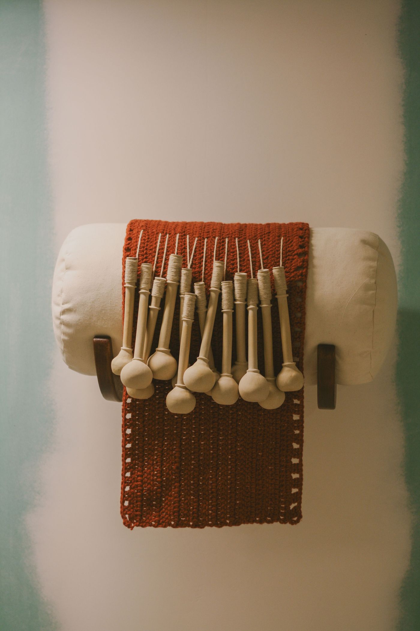 A wall-mounted art piece from the "Crafting with AI" exhibition at Estrela 74 features an array of wooden, elongated bobbin-like forms. They are neatly arranged on a vibrant red, open-weave textile, which is draped over a white, cylindrical cushion. The installation emphasizes traditional craftsmanship merged with contemporary display, creating textural depth.