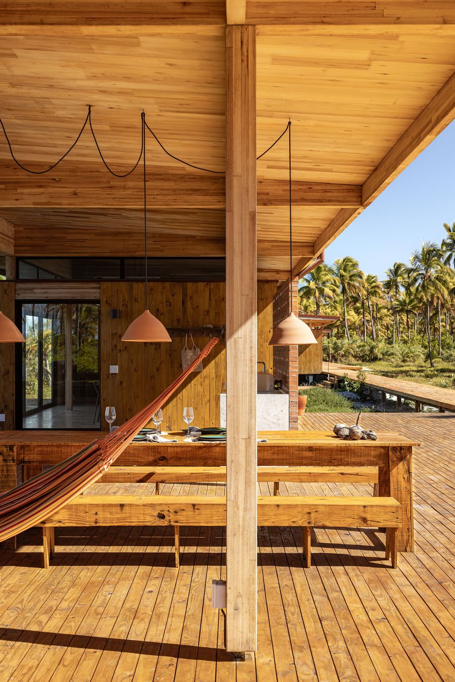An inviting outdoor dining space on a wooden deck features a large timber table and benches, with a hammock slung between supports. Overhanging ceramic pendants and the lush palm-lined backdrop create a relaxed, biophilic atmosphere, ideal for leisurely meals. An inviting outdoor dining space on a wooden deck features a large timber table and benches, with a hammock slung between supports. Overhanging ceramic pendants and the lush palm-lined backdrop create a relaxed, biophilic atmosphere, ideal for leisurely meals.