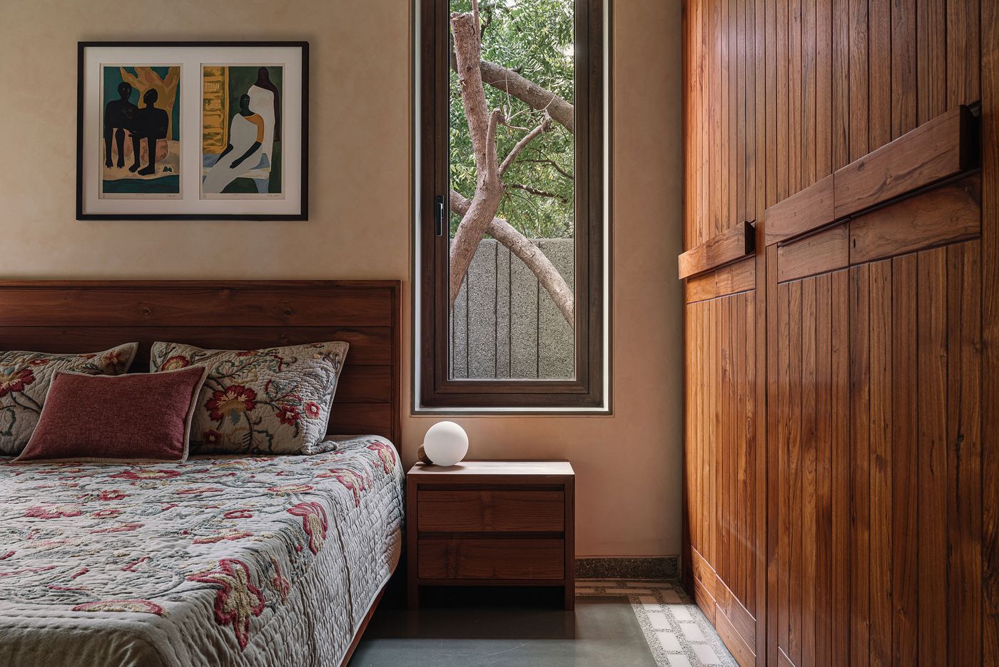 A warm and inviting bedroom with a rich wooden bed frame and complementary built-in wardrobe. The walls are a soft, earthy tone, providing a calm backdrop for a window framing a view of tree branches. A globe-shaped lamp on the bedside table and framed artwork featuring abstract figures add personal touches to this serene space.