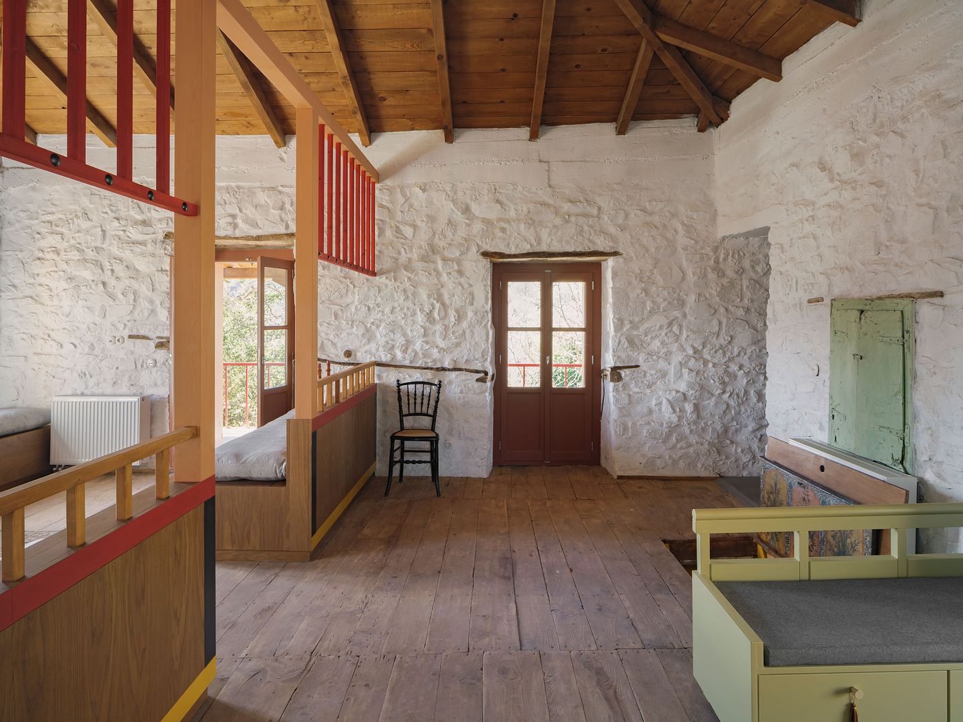 Wide-plank oak floors anchor this modular living arrangement where coral-red steel mezzanines create spatial hierarchy within the whitewashed stone interior. The traditional timber roof structure meets contemporary built-in furniture, demonstrating how neo-vernacular restoration balances historical authenticity with modern communal living requirements.
