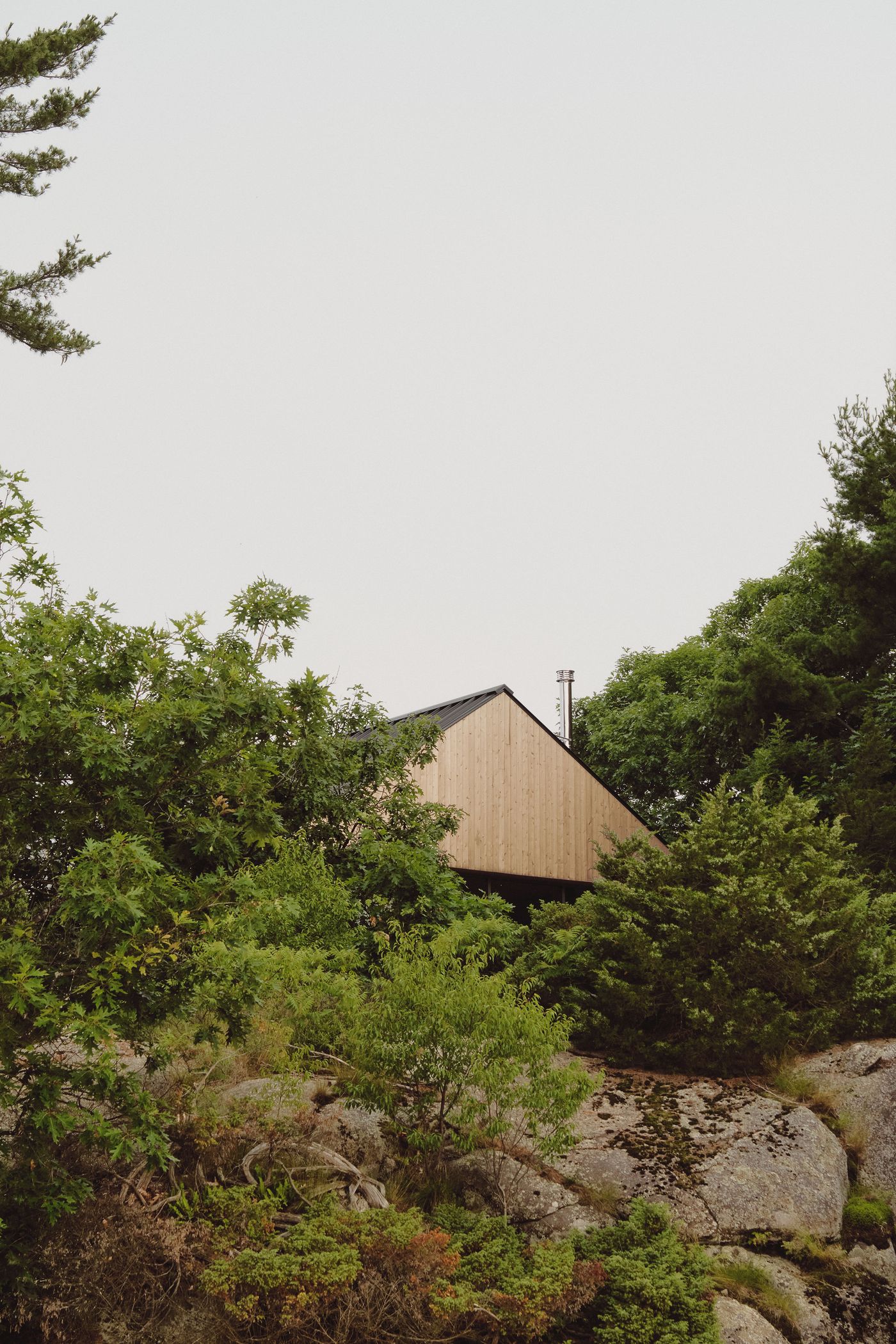 A modern wooden house with a triangular roof, nestled among lush greenery and rocky terrain, under a pale sky.
