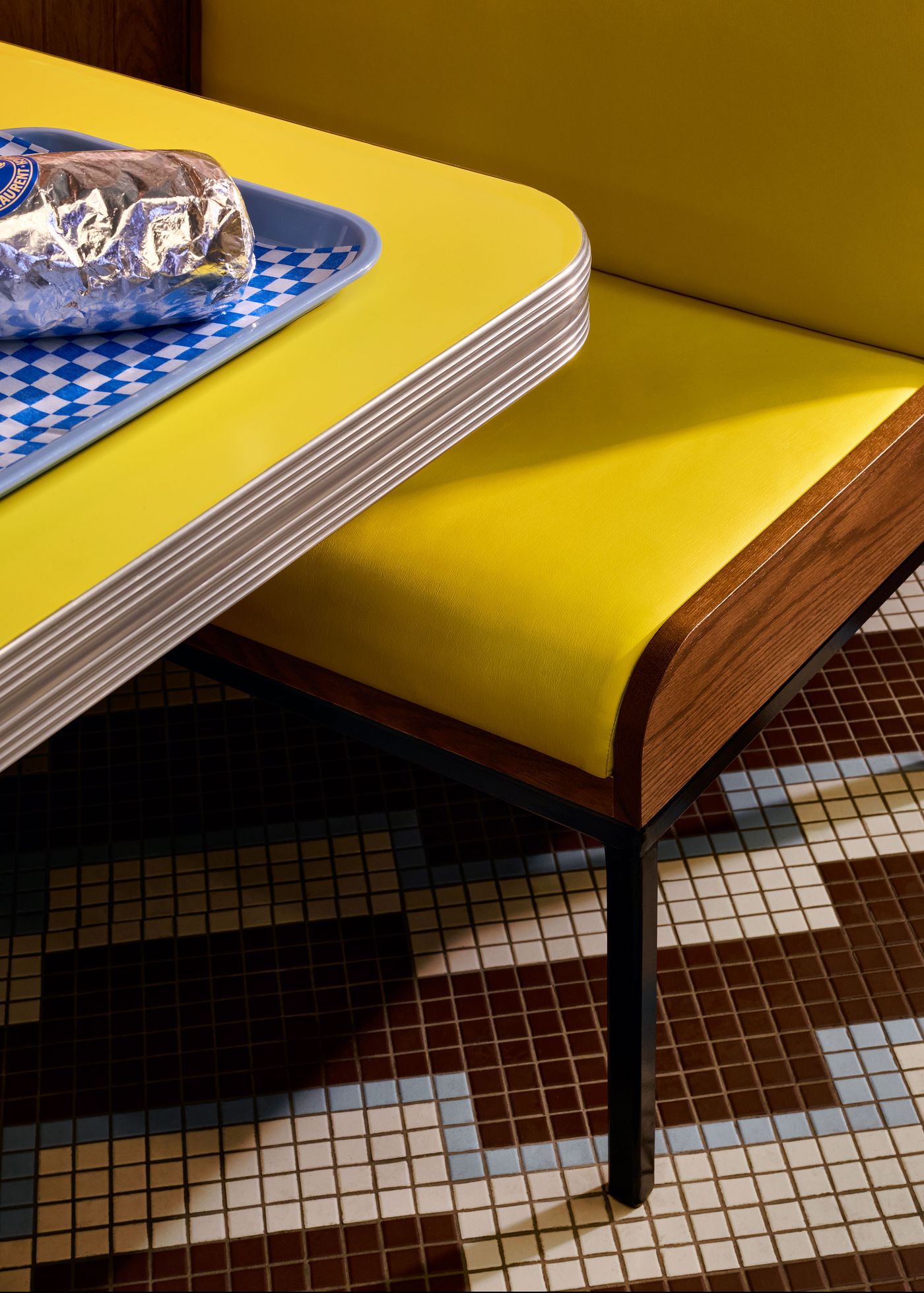 A close-up of a yellow diner table with a chrome trim, showcasing a sandwich wrapped in foil on a blue tray with a checkered pattern. The detail highlights the clean lines and materials of the furniture, emphasizing the retro design of the space.