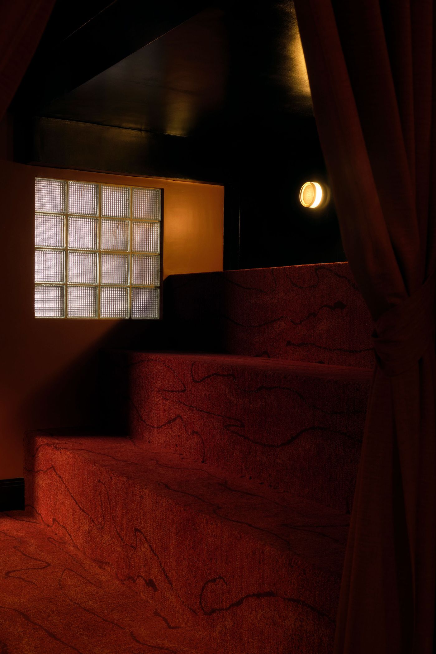 A dramatic, dimly lit interior staircase at The Roses of Elagabalus, covered in a plush, patterned red carpet that evokes a luxurious, vintage atmosphere. A grid of translucent glass blocks offers a soft, diffused light source, contrasting with the dark walls and single circular wall sconce, contributing to the space's secretive and opulent character.