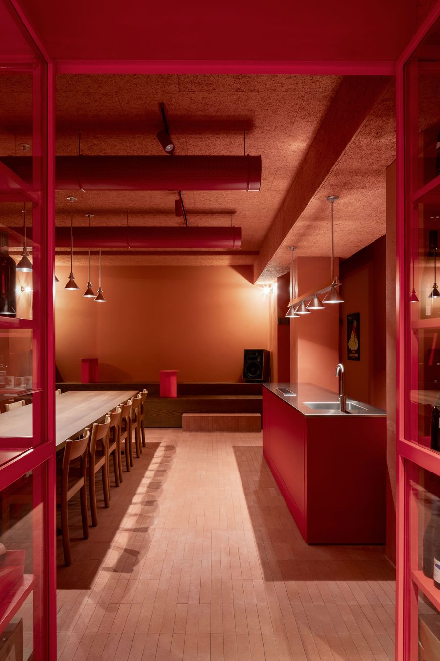 A versatile event space at Santo Palato by Naessi Studio, defined by a warm terracotta palette. A long wooden communal table and tiered wooden seating offer flexible arrangements. A vibrant red bar counter with a sink anchors one side, while exposed red ceiling ducts and minimalist lighting create a contemporary, industrial-chic ambiance. A versatile event space at Santo Palato by Naessi Studio, defined by a warm terracotta palette. A long wooden communal table and tiered wooden seating offer flexible arrangements. A vibrant red bar counter with a sink anchors one side, while exposed red ceiling ducts and minimalist lighting create a contemporary, industrial-chic ambiance.