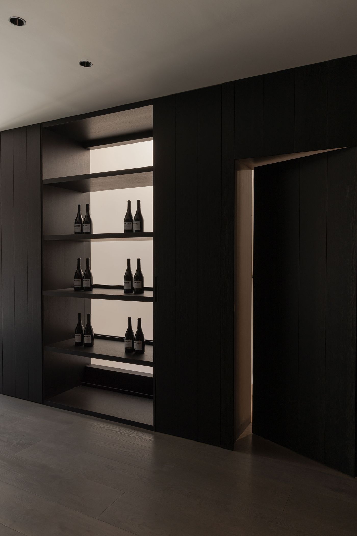 A sleek, dark wooden wall features illuminated shelves displaying wine bottles, with a slim door on the right leading to another space.
