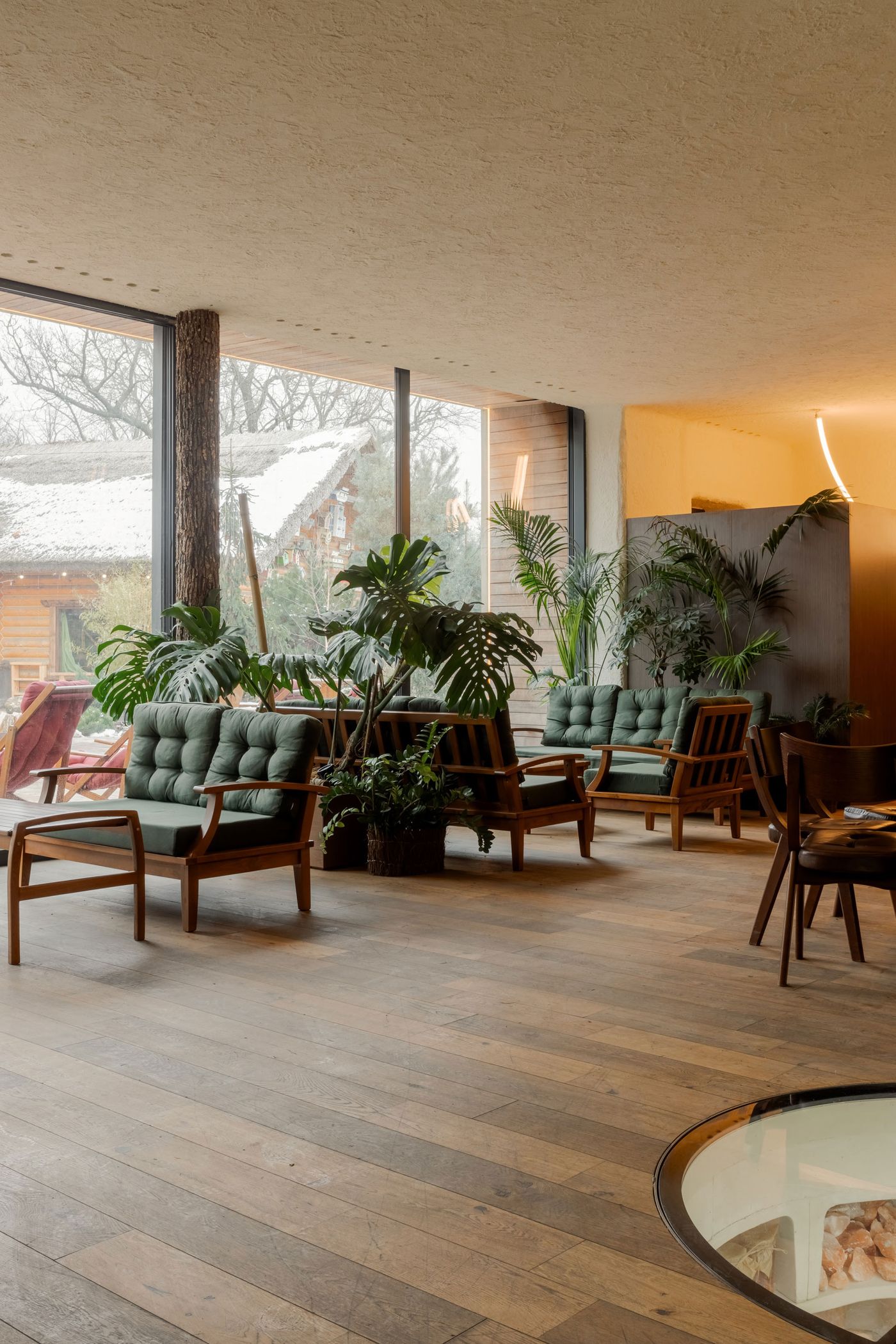 Cozy interior featuring green cushioned chairs, potted plants, and large windows with a snowy view outside. Warm wooden floors throughout. Cozy interior featuring green cushioned chairs, potted plants, and large windows with a snowy view outside. Warm wooden floors throughout.