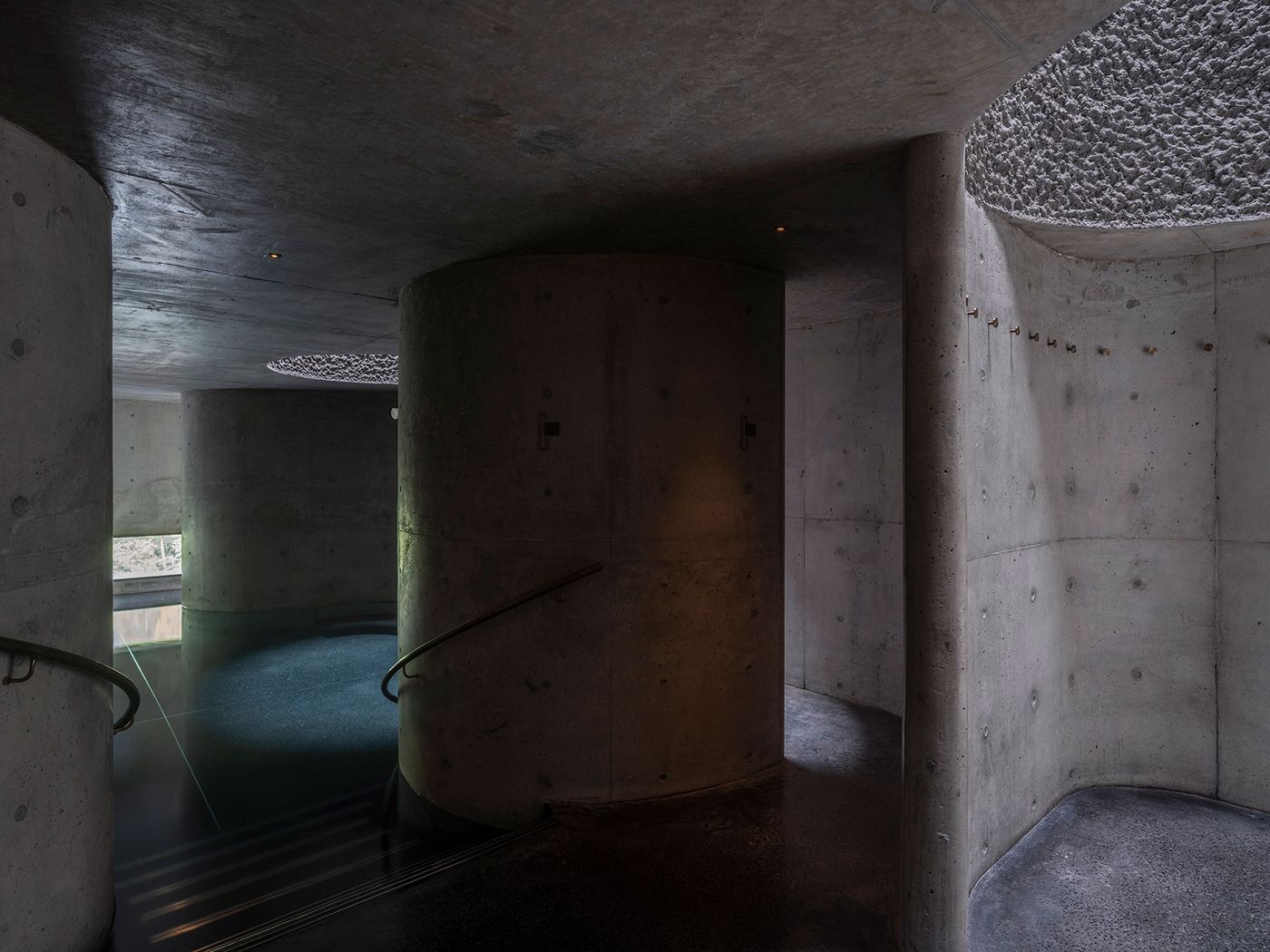 A dimly lit modern interior featuring curved concrete columns, a polished floor, and soft light filtering through an overhead opening. A dimly lit modern interior featuring curved concrete columns, a polished floor, and soft light filtering through an overhead opening.