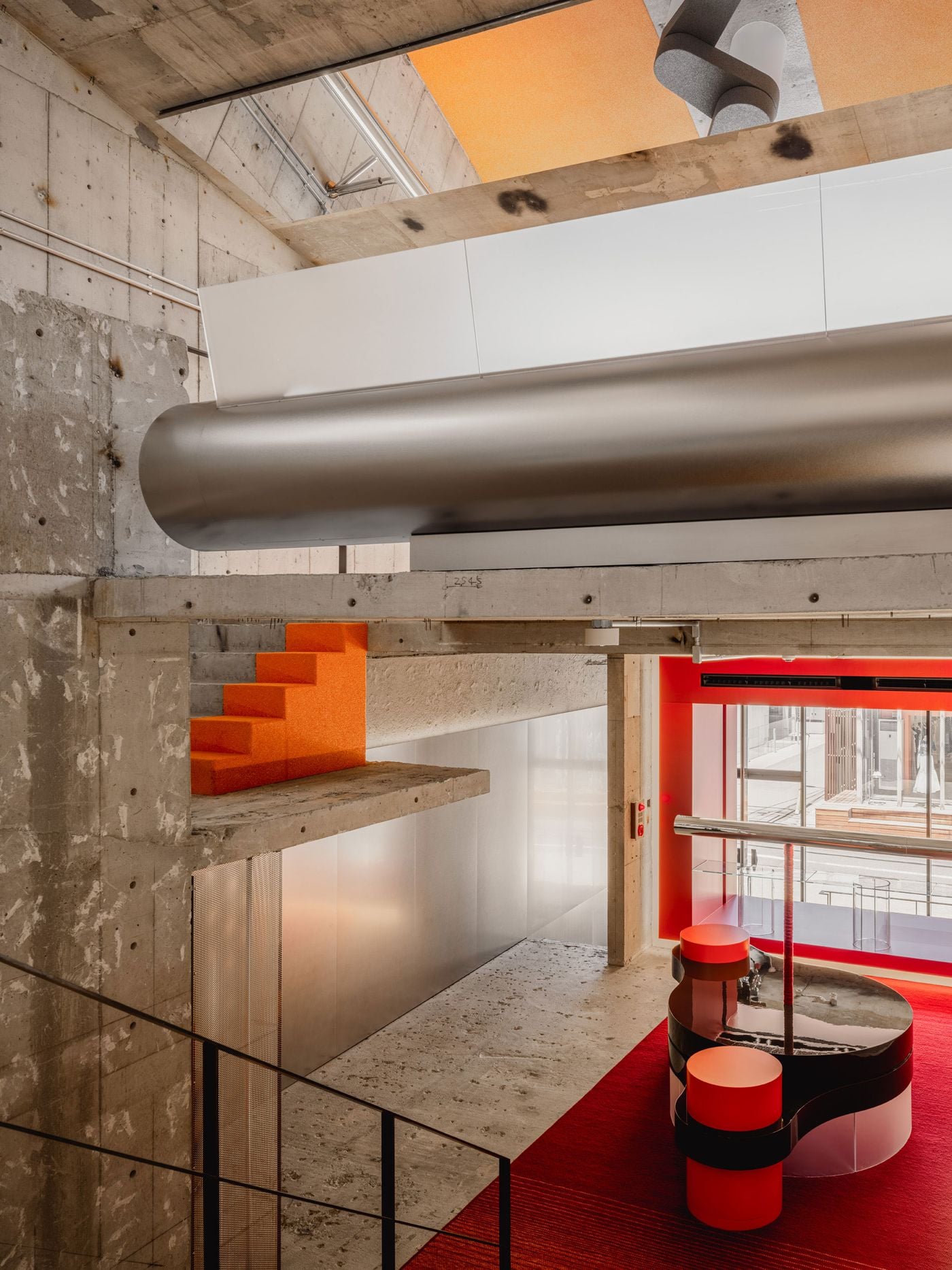 Multiple levels overlap in this view through the store, where a large brushed-metal cylindrical volume cuts horizontally across the exposed concrete structure. A small flight of orange steps punctuates the composition, while the red-framed ground floor remains visible below, emphasising the project’s vertical continuity and contrasting material palette.