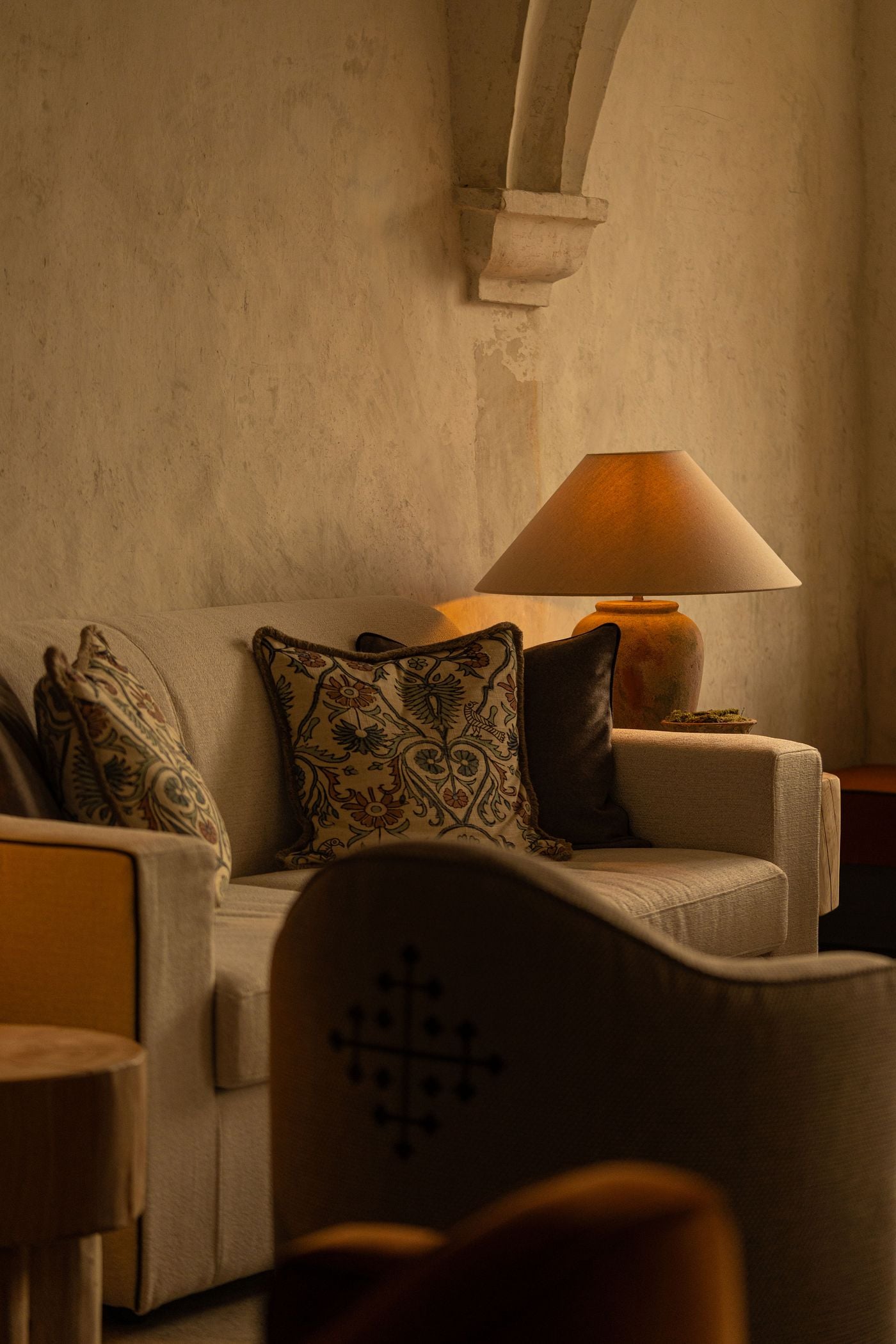 A vault rib springs from a carved stone corbel above a cream sofa at Castel Badia, set against softly mottled plaster. Fringed cushions embroidered with suzani-style florals, a terracotta lamp with a linen shade and the cross-stitched back of an armchair in the foreground compose a quiet, tonal vignette. A vault rib springs from a carved stone corbel above a cream sofa at Castel Badia, set against softly mottled plaster. Fringed cushions embroidered with suzani-style florals, a terracotta lamp with a linen shade and the cross-stitched back of an armchair in the foreground compose a quiet, tonal vignette.
