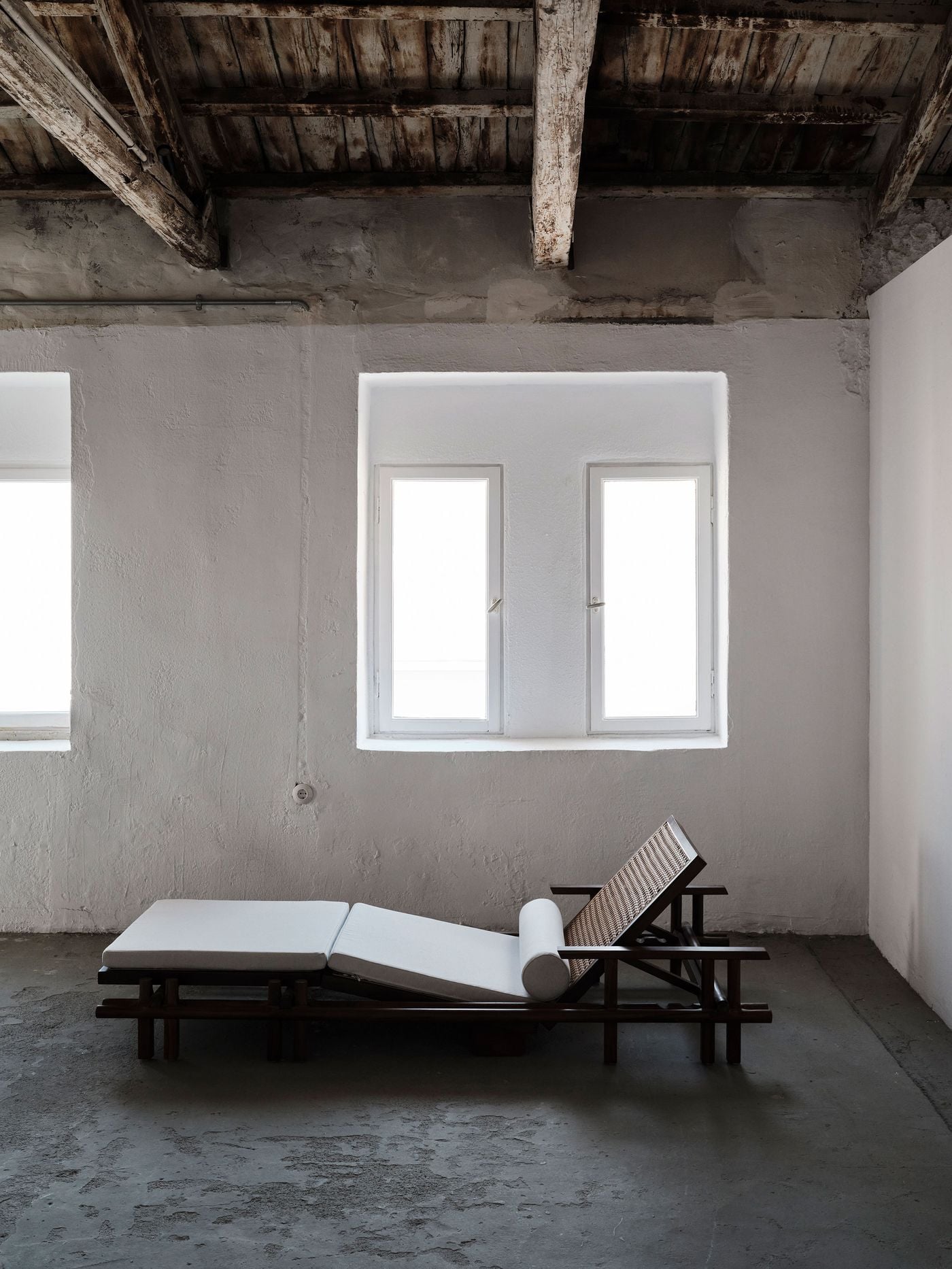A low timber-and-cane daybed anchors a sparse upper-floor room beneath the original weathered timber roof. Whitewashed textured walls and deep-set windows amplify the natural light, while the worn concrete floor and exposed roof structure preserve the building’s raw, timeworn character. A low timber-and-cane daybed anchors a sparse upper-floor room beneath the original weathered timber roof. Whitewashed textured walls and deep-set windows amplify the natural light, while the worn concrete floor and exposed roof structure preserve the building’s raw, timeworn character.