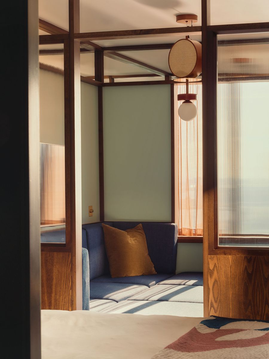 The blue sofa pit is glimpsed through a framework of dark walnut and reeded glass, illustrating how Holloway Li divides the guest room into distinct yet visually connected zones. Sage-green walls, rust-toned curtains and warm afternoon light soften the strong geometry and deepen the mid-century atmosphere. The blue sofa pit is glimpsed through a framework of dark walnut and reeded glass, illustrating how Holloway Li divides the guest room into distinct yet visually connected zones. Sage-green walls, rust-toned curtains and warm afternoon light soften the strong geometry and deepen the mid-century atmosphere.