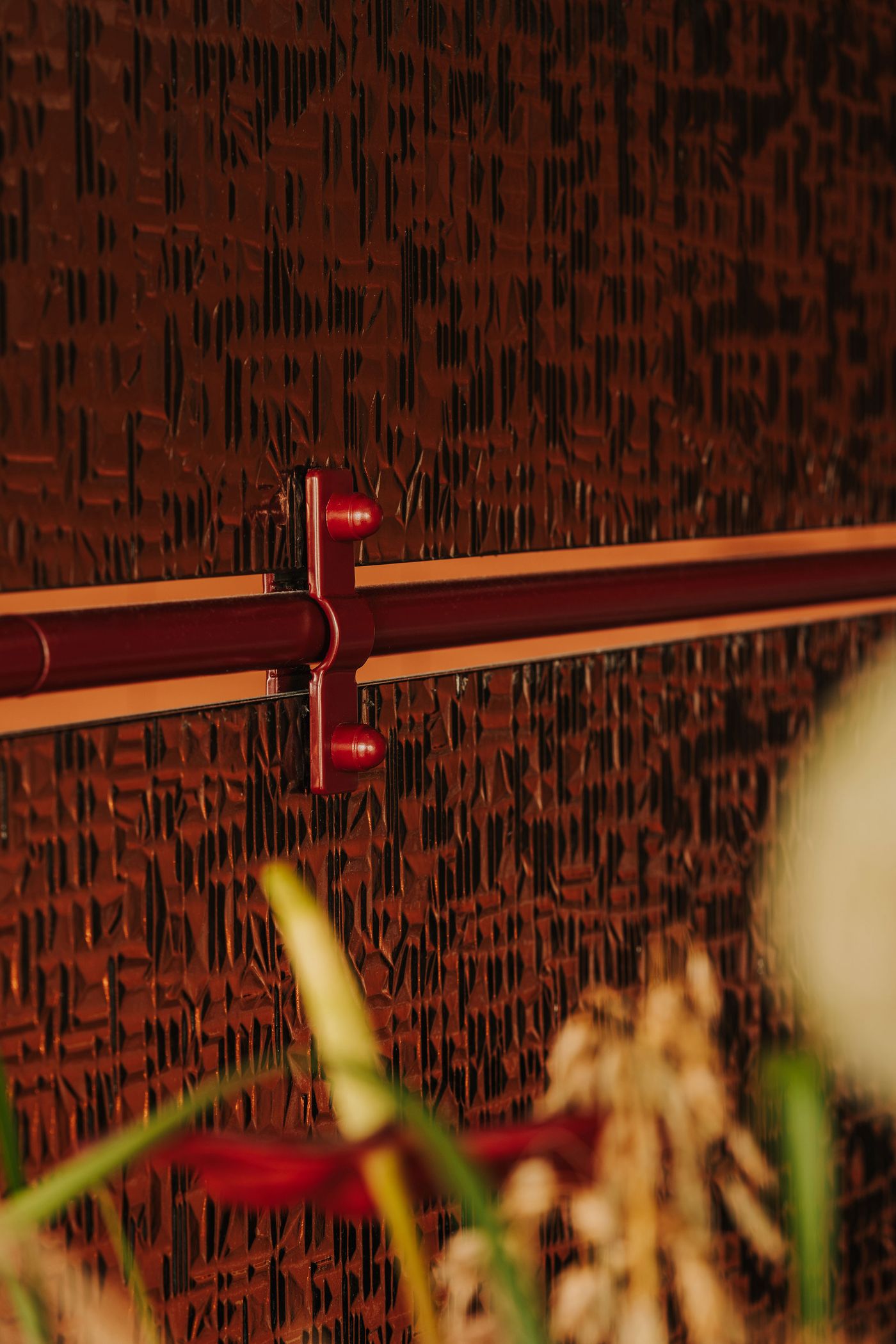 An extreme close-up of WIN’s ornamental glass partition reveals its deeply textured, faceted surface, framed by burgundy metal tubing and small matching brackets. The interplay of light and shadow accentuates the glass’s irregular relief, highlighting the tactile detailing that softens the wine bar’s otherwise industrial aesthetic. An extreme close-up of WIN’s ornamental glass partition reveals its deeply textured, faceted surface, framed by burgundy metal tubing and small matching brackets. The interplay of light and shadow accentuates the glass’s irregular relief, highlighting the tactile detailing that softens the wine bar’s otherwise industrial aesthetic.