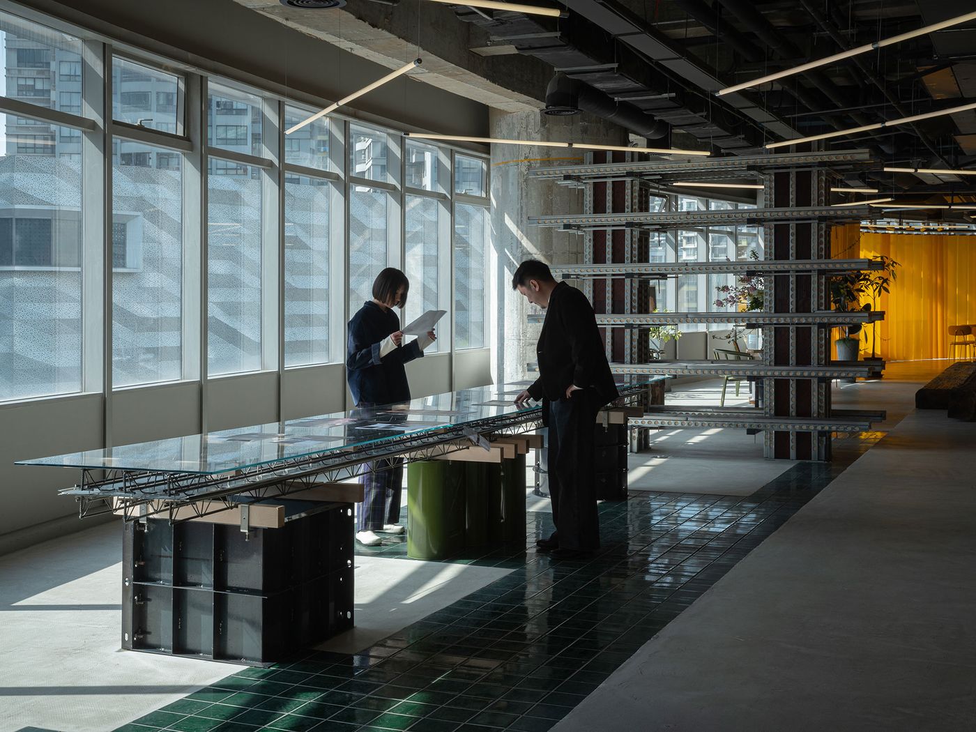 In 889 GLO Art Space, two individuals interact beside a long, custom-designed glass table, revealing intricate metalwork beneath. The space is flooded with natural light from large windows, highlighting the striking contrast between the industrial aesthetic of exposed structures, a dark green tiled floor, and the vibrant yellow partitions in the background. In 889 GLO Art Space, two individuals interact beside a long, custom-designed glass table, revealing intricate metalwork beneath. The space is flooded with natural light from large windows, highlighting the striking contrast between the industrial aesthetic of exposed structures, a dark green tiled floor, and the vibrant yellow partitions in the background.