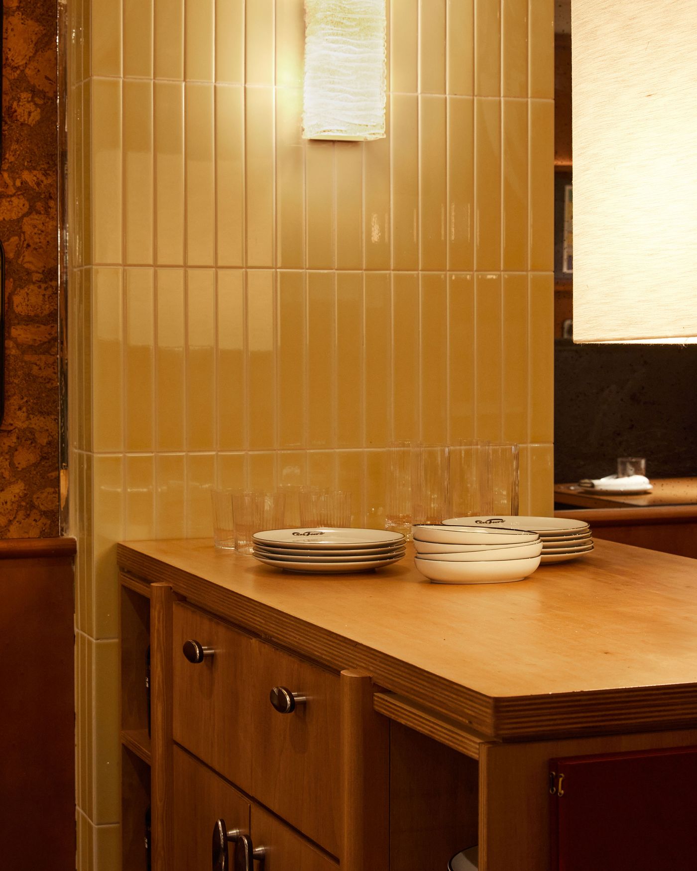 A detailed shot of a custom-built counter, showcasing a thoughtful material palette. A column covered in narrow, cream-colored tiles and a textured glass light fixture creates a striking vertical line, while the warm wooden surface of the counter adds a natural, grounding element to the retro-inspired design.