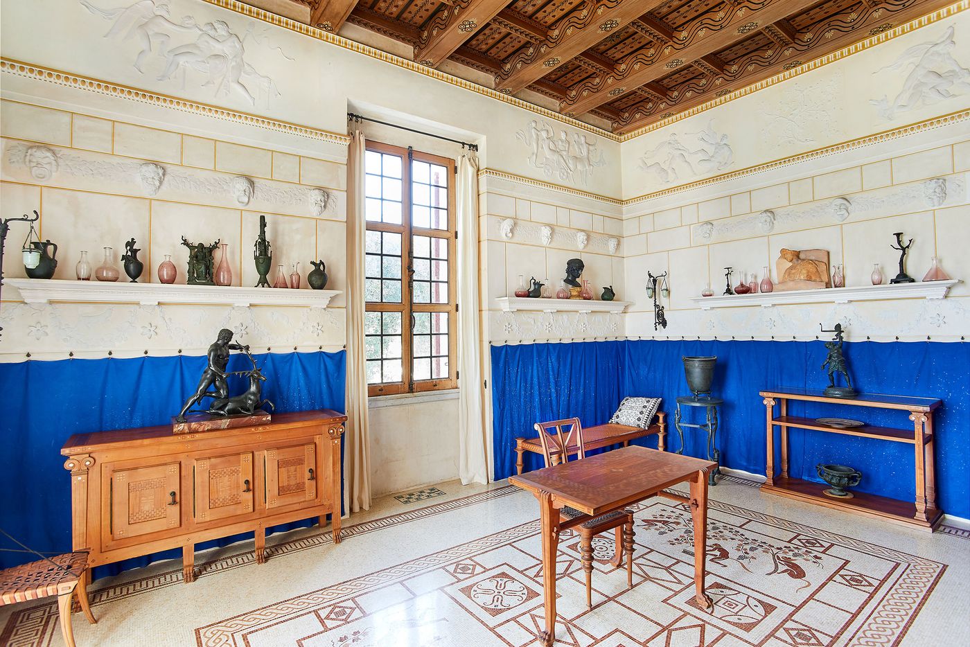 An interior room at Villa Kerylos with a blend of classical and modern aesthetics. The space features a richly textured coffered ceiling and a mosaic tile floor. A bright blue curtain adds a bold splash of color against the pale walls with their white bas-reliefs, while antique bronze objects are displayed on minimalist white ledges. An interior room at Villa Kerylos with a blend of classical and modern aesthetics. The space features a richly textured coffered ceiling and a mosaic tile floor. A bright blue curtain adds a bold splash of color against the pale walls with their white bas-reliefs, while antique bronze objects are displayed on minimalist white ledges.