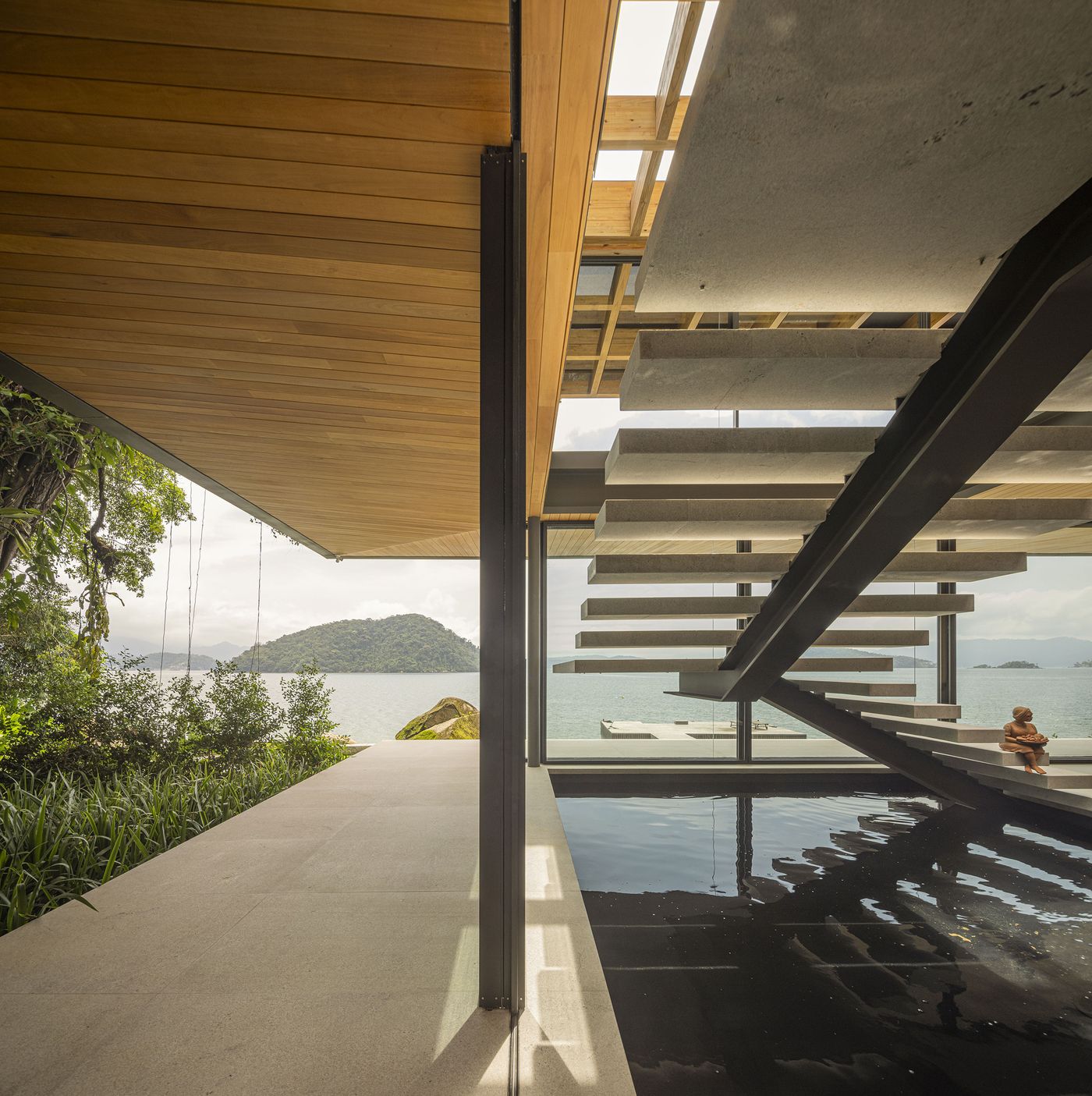 This striking image captures an indoor-outdoor swimming pool seamlessly merging with the horizon, offering expansive views of the ocean and distant islands. An architectural staircase with floating concrete treads creates a dramatic vertical element against the light-filled, minimalist space, while a figure relaxes poolside, emphasizing tranquility. This striking image captures an indoor-outdoor swimming pool seamlessly merging with the horizon, offering expansive views of the ocean and distant islands. An architectural staircase with floating concrete treads creates a dramatic vertical element against the light-filled, minimalist space, while a figure relaxes poolside, emphasizing tranquility.