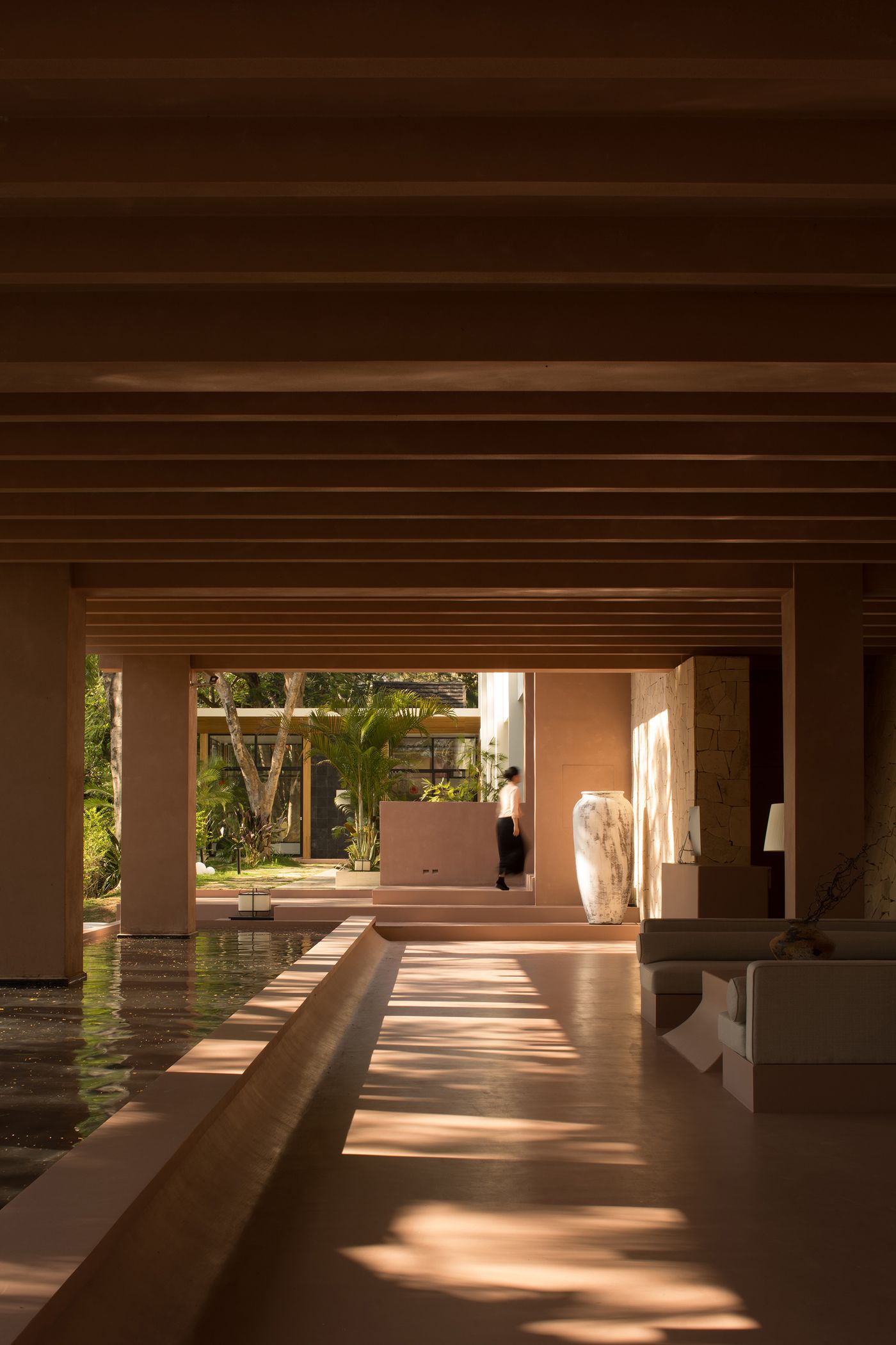 Interior view of a modern, minimalist space with warm tones, featuring a water feature, lush greenery, and a person walking by. Interior view of a modern, minimalist space with warm tones, featuring a water feature, lush greenery, and a person walking by.