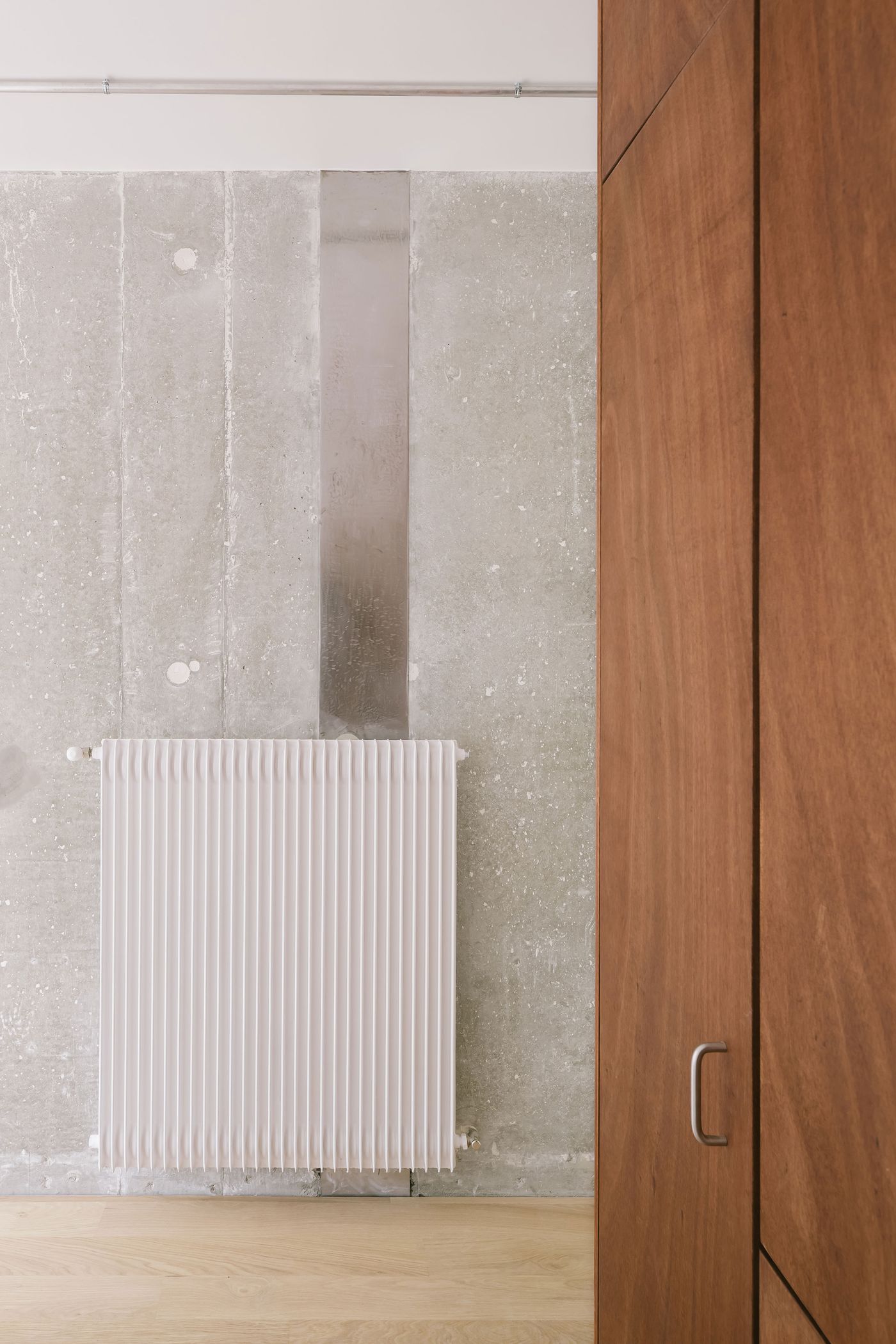 Close-up of a white radiator against a textured concrete wall, with a wooden wardrobe edge visible on the right side.