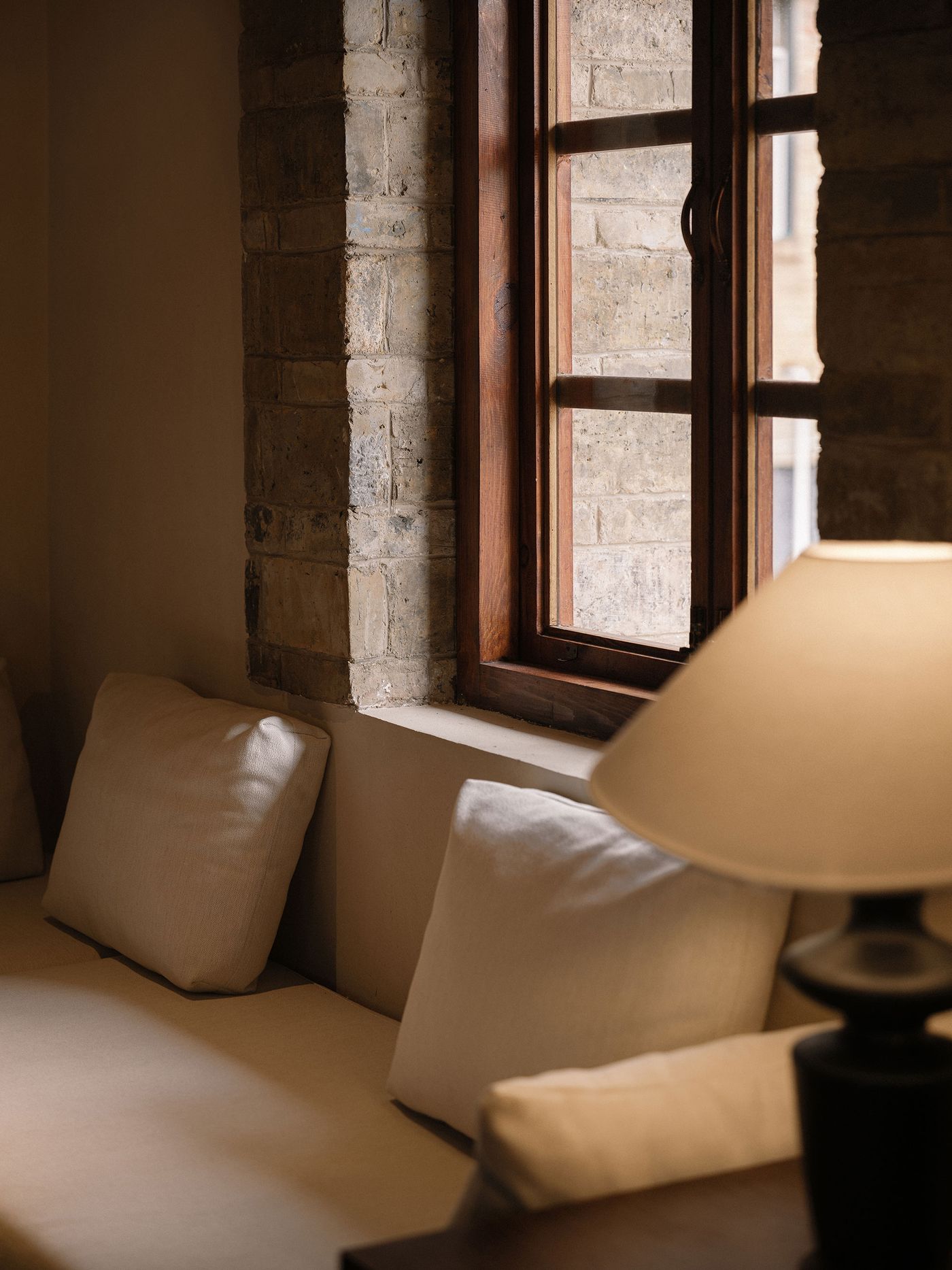A cozy corner with beige pillows on a sofa, a stone wall, wooden window frames, and a soft lamp illuminating the space.
