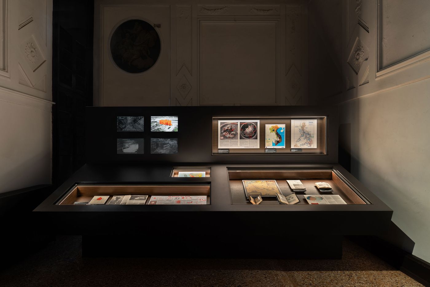 A dark exhibition space featuring a display case with maps, documents, and digital panels presenting historical imagery and data. A dark exhibition space featuring a display case with maps, documents, and digital panels presenting historical imagery and data.
