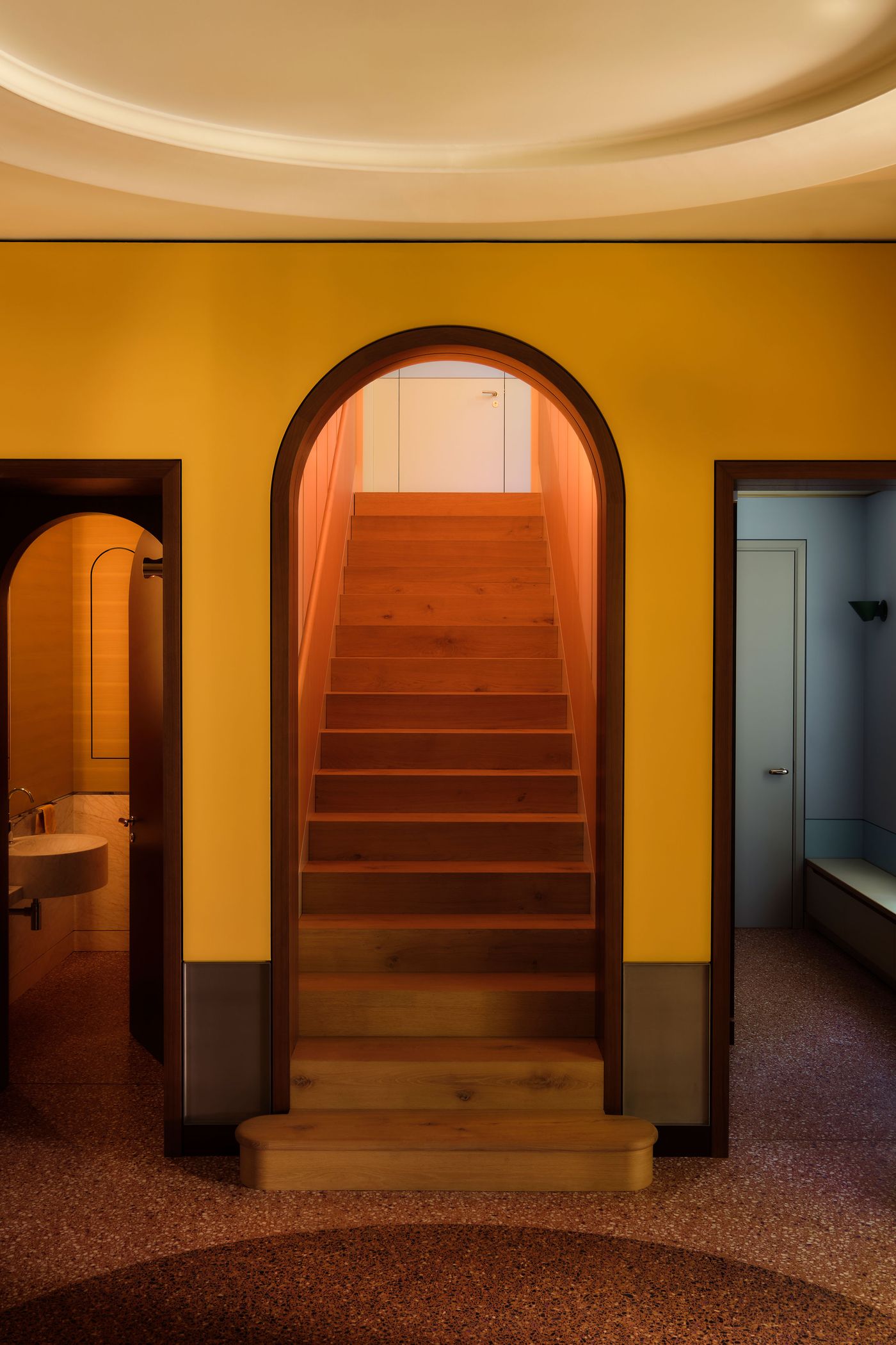 Brightly lit hallway featuring wooden stairs, yellow walls, and arched doorways leading to a bathroom and another room.