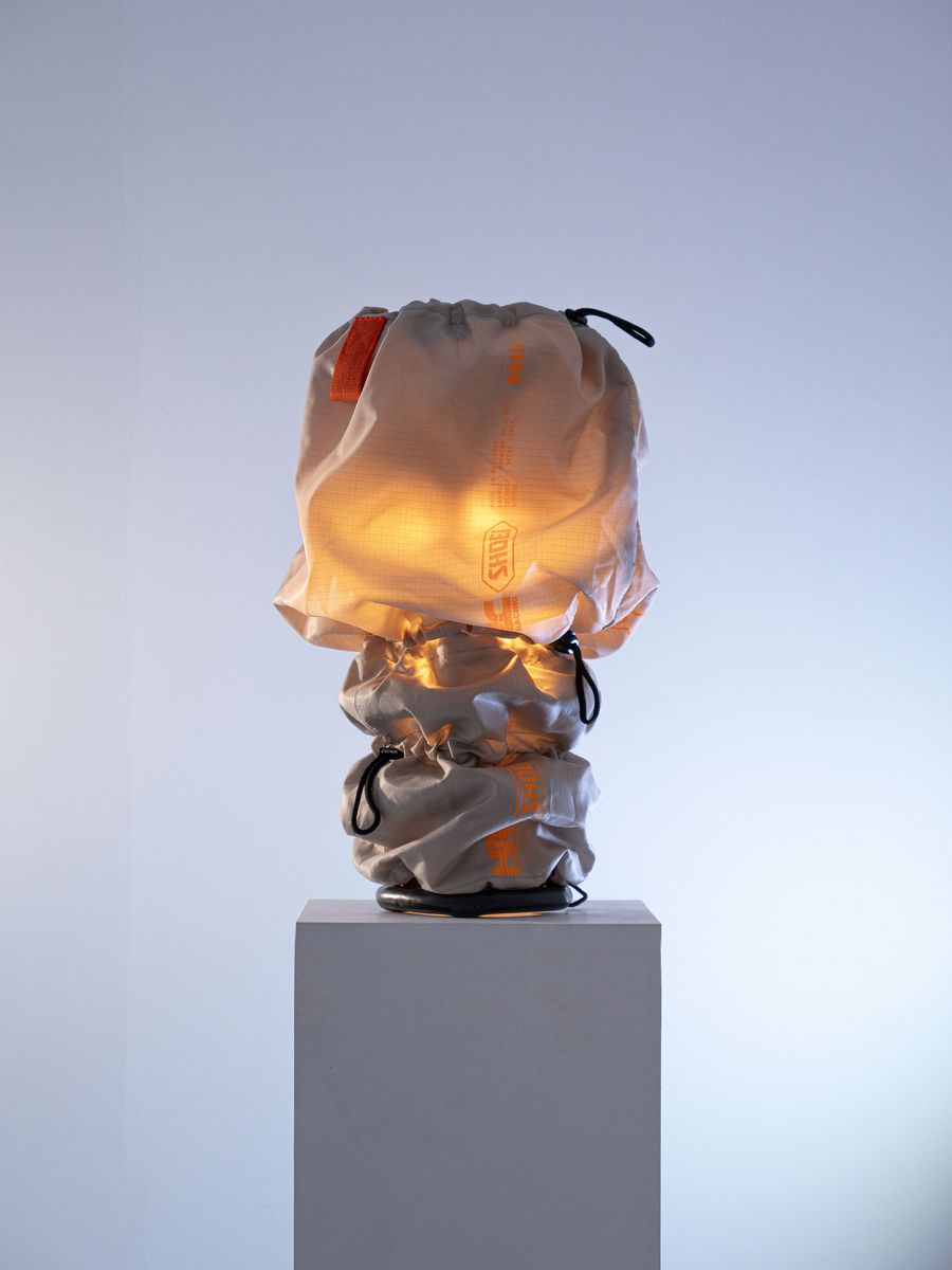 A unique lamp covered in a translucent, drawstring fabric emits a warm glow, displayed atop a simple white pedestal against a soft backdrop. A unique lamp covered in a translucent, drawstring fabric emits a warm glow, displayed atop a simple white pedestal against a soft backdrop.