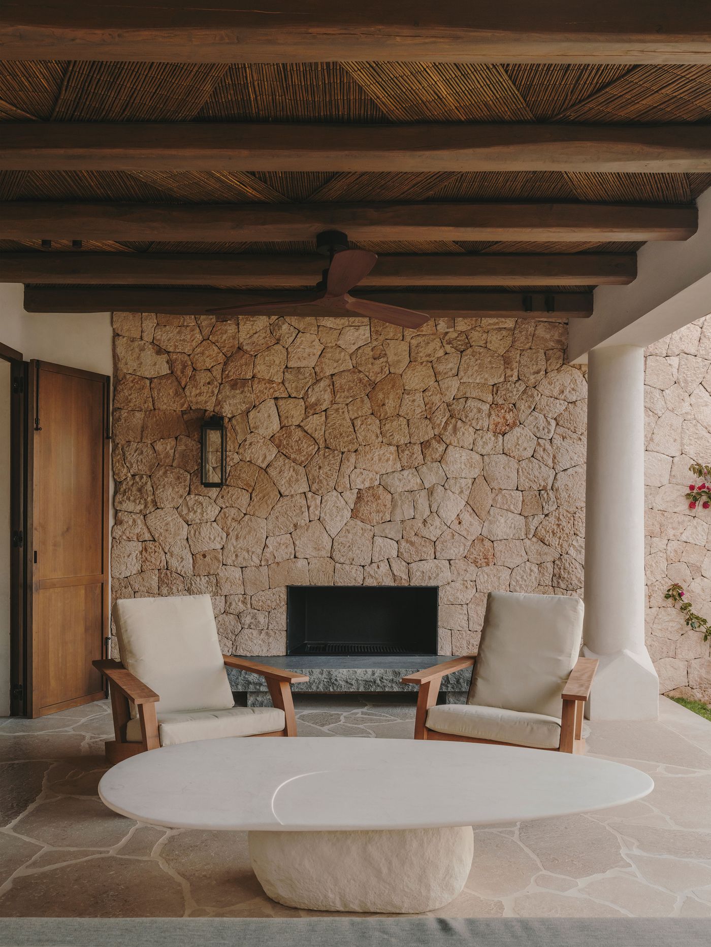 A stately outdoor seating area featuring two light-colored armchairs, a rounded white table, wooden roof beams and a stone wall backdrop.