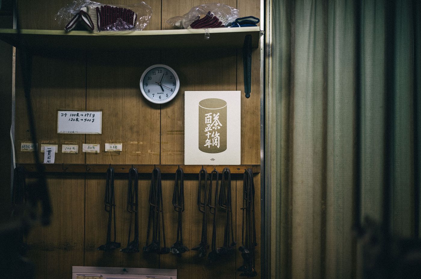 A wall featuring a clock, hanging tools, and a poster with Japanese text, set against a backdrop of curtains in a dimly lit room. A wall featuring a clock, hanging tools, and a poster with Japanese text, set against a backdrop of curtains in a dimly lit room.