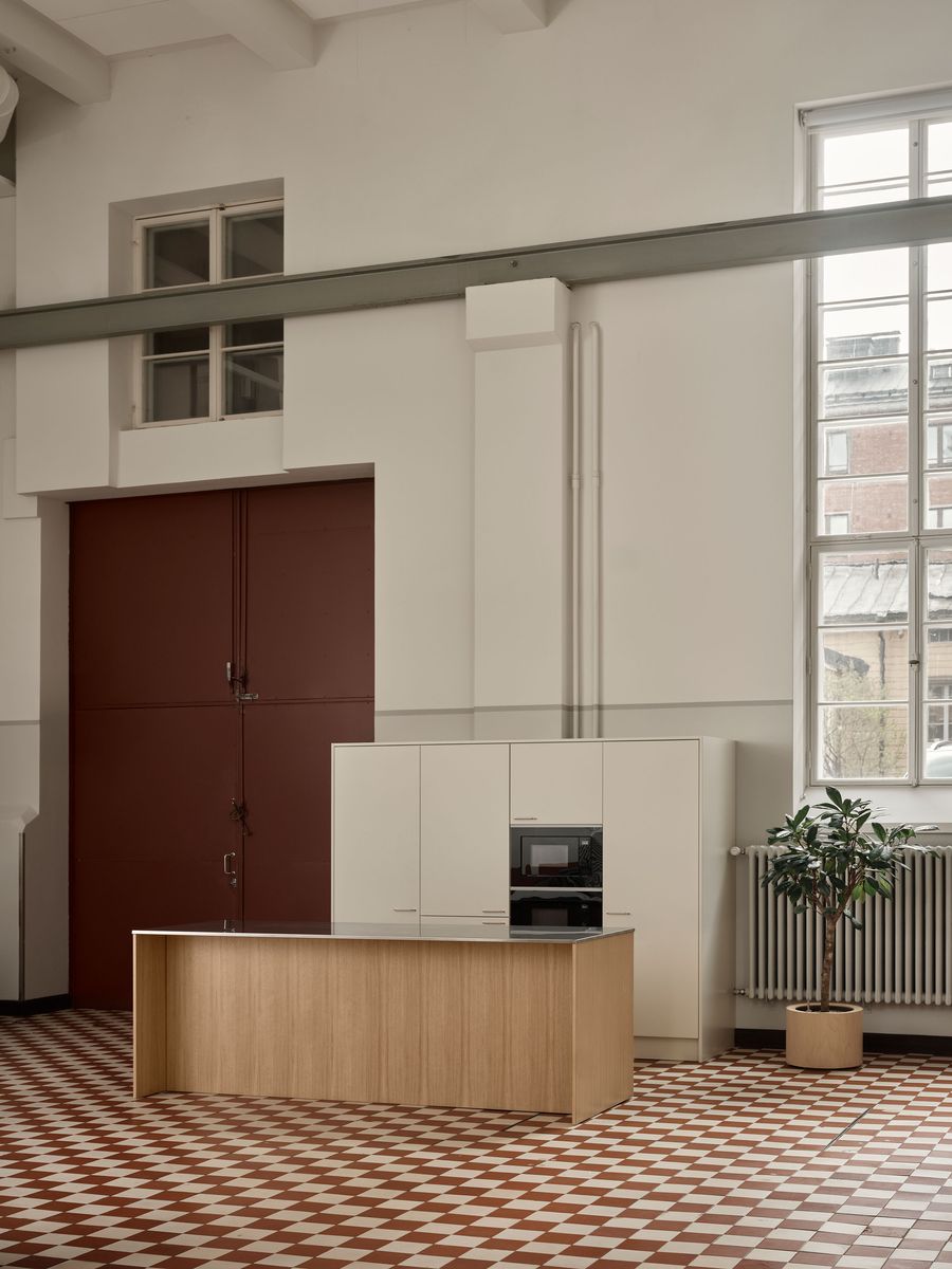 A Scandinavian industrial-chic kitchen within Konelabra, Helsinki by Fyra, featuring a bold red industrial door contrasting with the neutral walls. A light wood island and cream-colored minimalist cabinetry stand on a distinctive red and white checkerboard tile floor. Tall, multi-paned windows illuminate the space, highlighting the robust ceiling beams and an elegant potted plant. A Scandinavian industrial-chic kitchen within Konelabra, Helsinki by Fyra, featuring a bold red industrial door contrasting with the neutral walls. A light wood island and cream-colored minimalist cabinetry stand on a distinctive red and white checkerboard tile floor. Tall, multi-paned windows illuminate the space, highlighting the robust ceiling beams and an elegant potted plant.