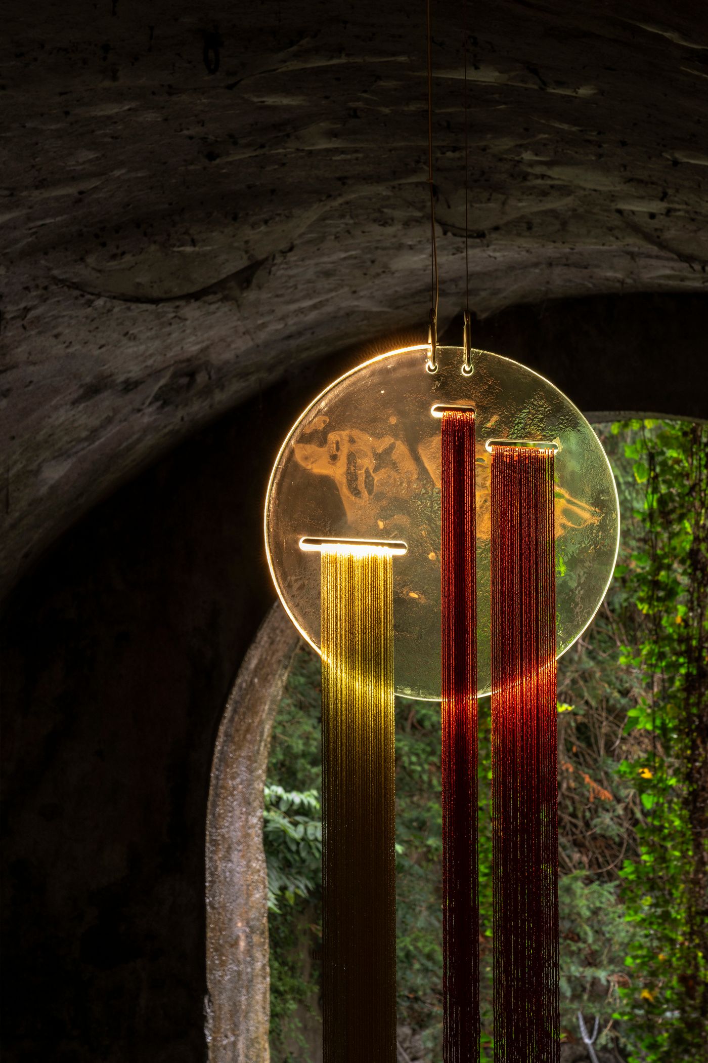 A circular, translucent disc, illuminated from behind, features three strands of shimmering metallic fringe—gold on the left, and red on the right—cascading downwards. This delicate hanging installation is set within a dark, arched architectural space, with lush greenery visible in the background through an opening. The play of light and the linear elements create a mesmerizing, almost ethereal effect, suggesting a contemporary art piece.
