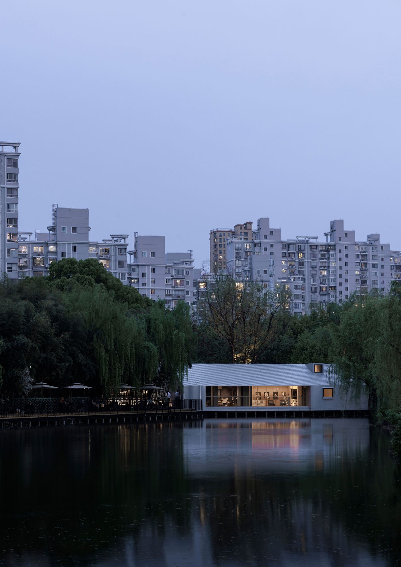 A striking minimalist structure, emerges from the twilight, its interior lights glowing warmly and reflecting on the calm lake. Towering urban buildings form a hazy backdrop, highlighting the secluded elegance of the house amidst the natural, verdant surroundings. The composition underscores the architectural design's ability to create an intimate sanctuary within an expansive city. A striking minimalist structure, emerges from the twilight, its interior lights glowing warmly and reflecting on the calm lake. Towering urban buildings form a hazy backdrop, highlighting the secluded elegance of the house amidst the natural, verdant surroundings. The composition underscores the architectural design's ability to create an intimate sanctuary within an expansive city.