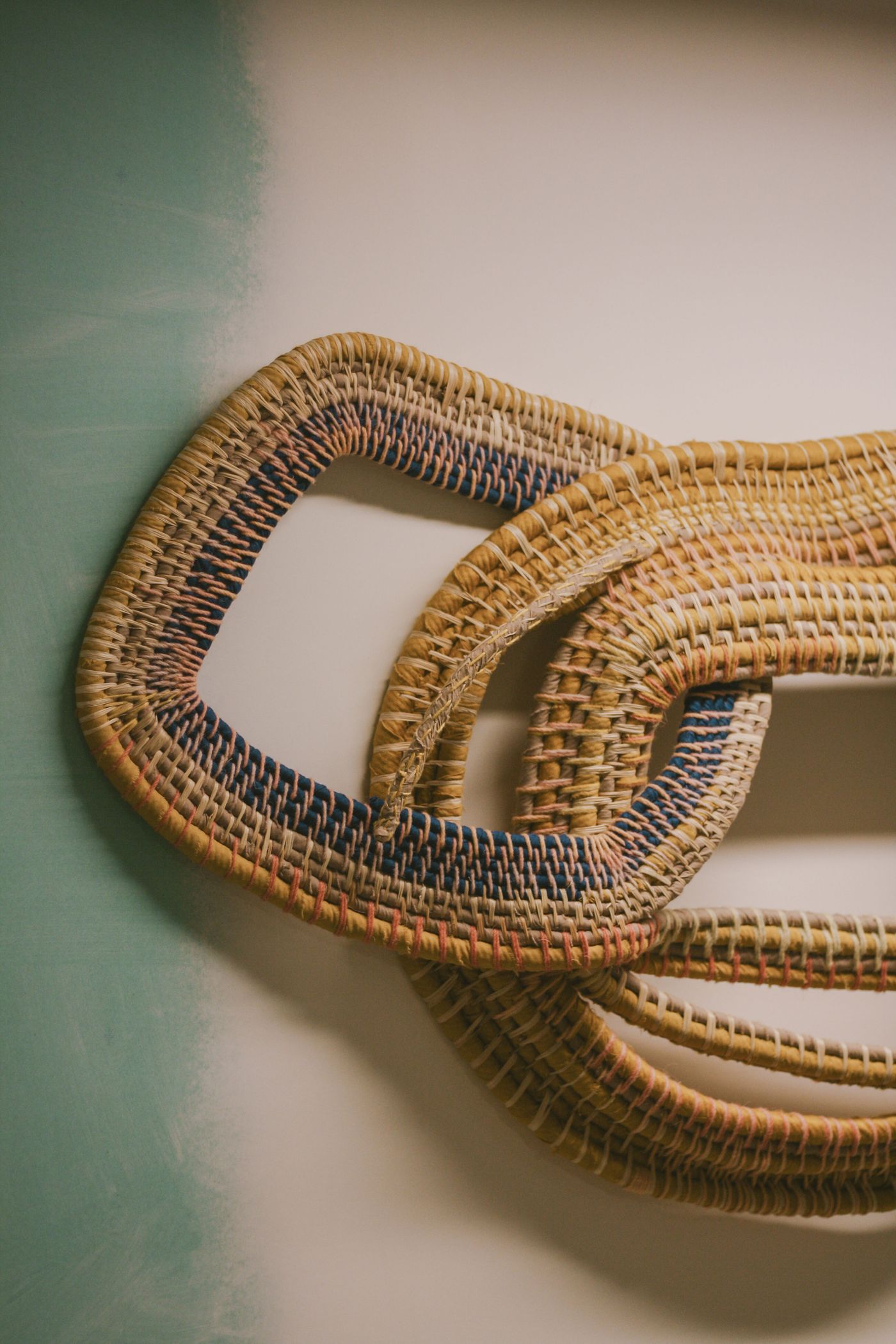 A close-up of a vibrant woven artwork from the "Crafting with AI" exhibition at Estrela 74. Interlocking, large-scale, sinuous forms crafted from straw or wicker feature intricate weaving with accents of blue, red, and yellow threads. The organic shapes and rich textures are set against a partially painted, raw teal and white wall, highlighting its artisanal quality.