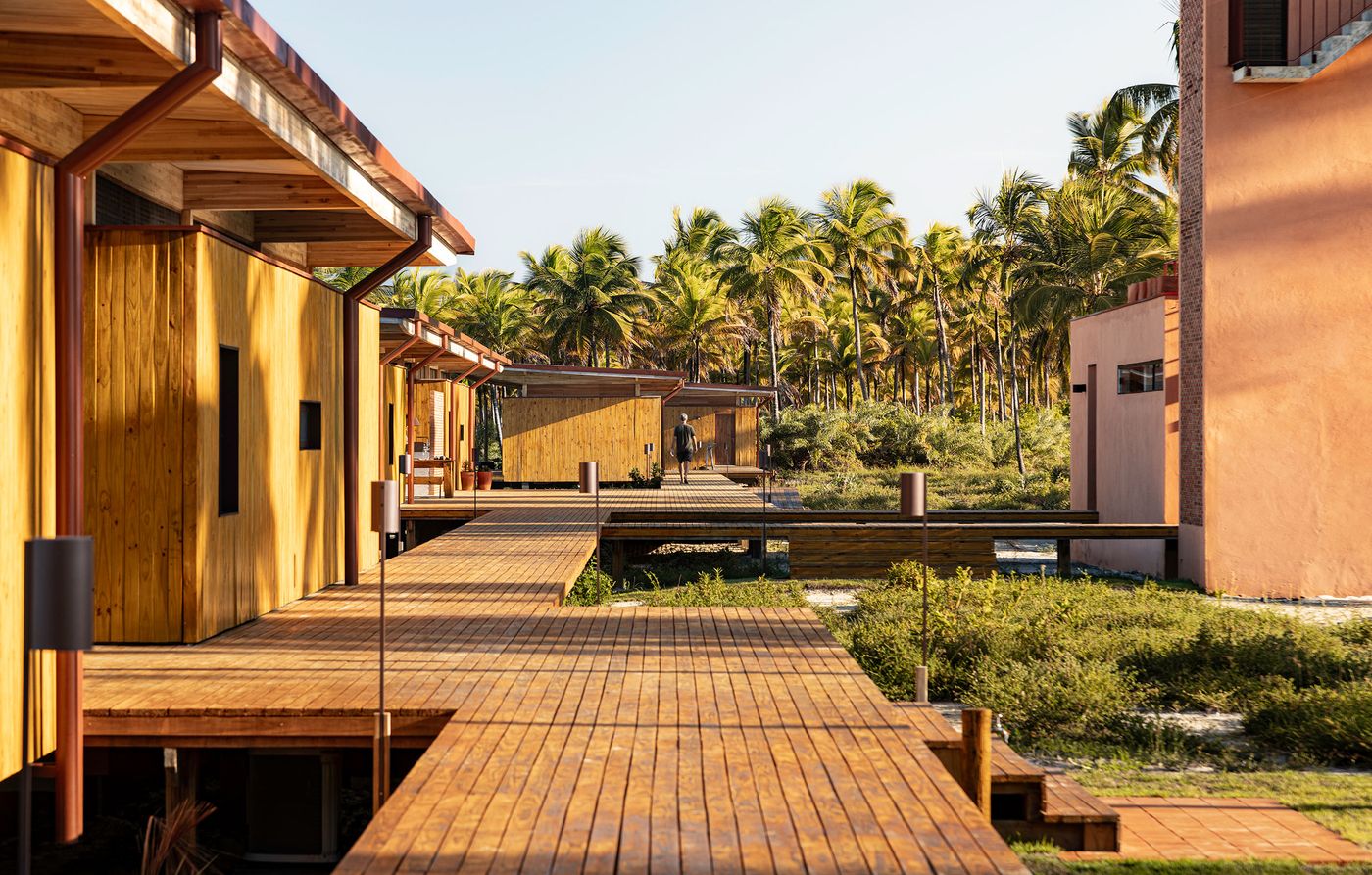A series of elevated wooden walkways connect modern structures with warm timber cladding and a distinct peach-toned building. Surrounded by vibrant tropical foliage and palm trees, the design emphasizes a seamless, integrated living experience within nature. A series of elevated wooden walkways connect modern structures with warm timber cladding and a distinct peach-toned building. Surrounded by vibrant tropical foliage and palm trees, the design emphasizes a seamless, integrated living experience within nature.