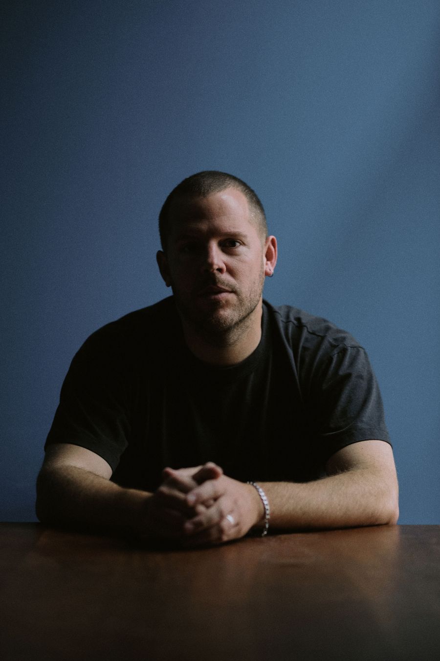 A person with cropped hair sits at a wooden table, clasping their hands, against a blue wall background. A person with cropped hair sits at a wooden table, clasping their hands, against a blue wall background.