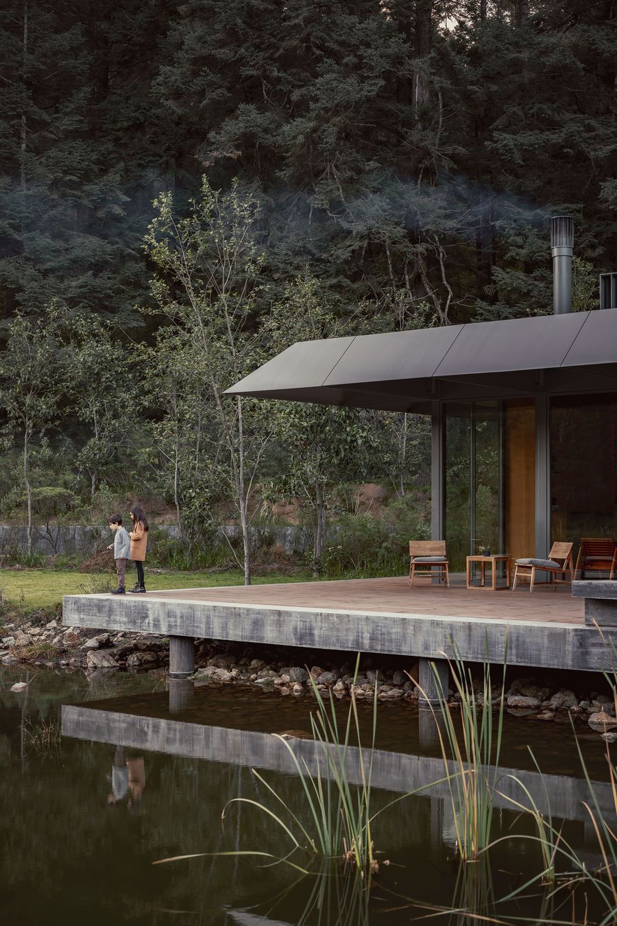 A girl and a boy stand by a serene lakeside cabin, surrounded by lush trees, with smoke gently rising from the chimney.