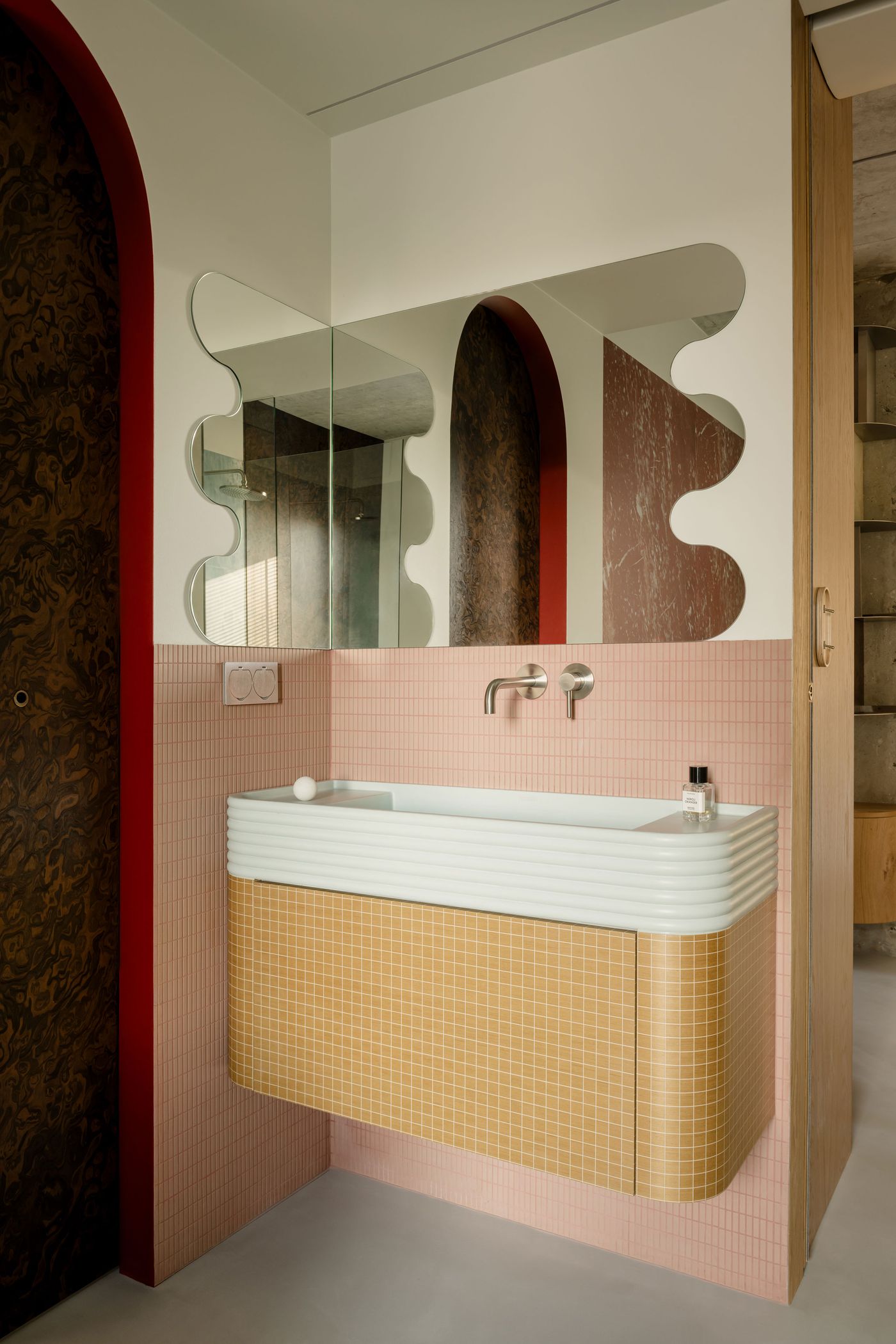 This bathroom composition balances retro curves and bold geometry, with a sculptural mirror, arched walnut niche, and a pastel basin fronted by mustard mosaic tiles. The contrast between red, brown, and soft pink defines the postmodern spirit of the space. This bathroom composition balances retro curves and bold geometry, with a sculptural mirror, arched walnut niche, and a pastel basin fronted by mustard mosaic tiles. The contrast between red, brown, and soft pink defines the postmodern spirit of the space.