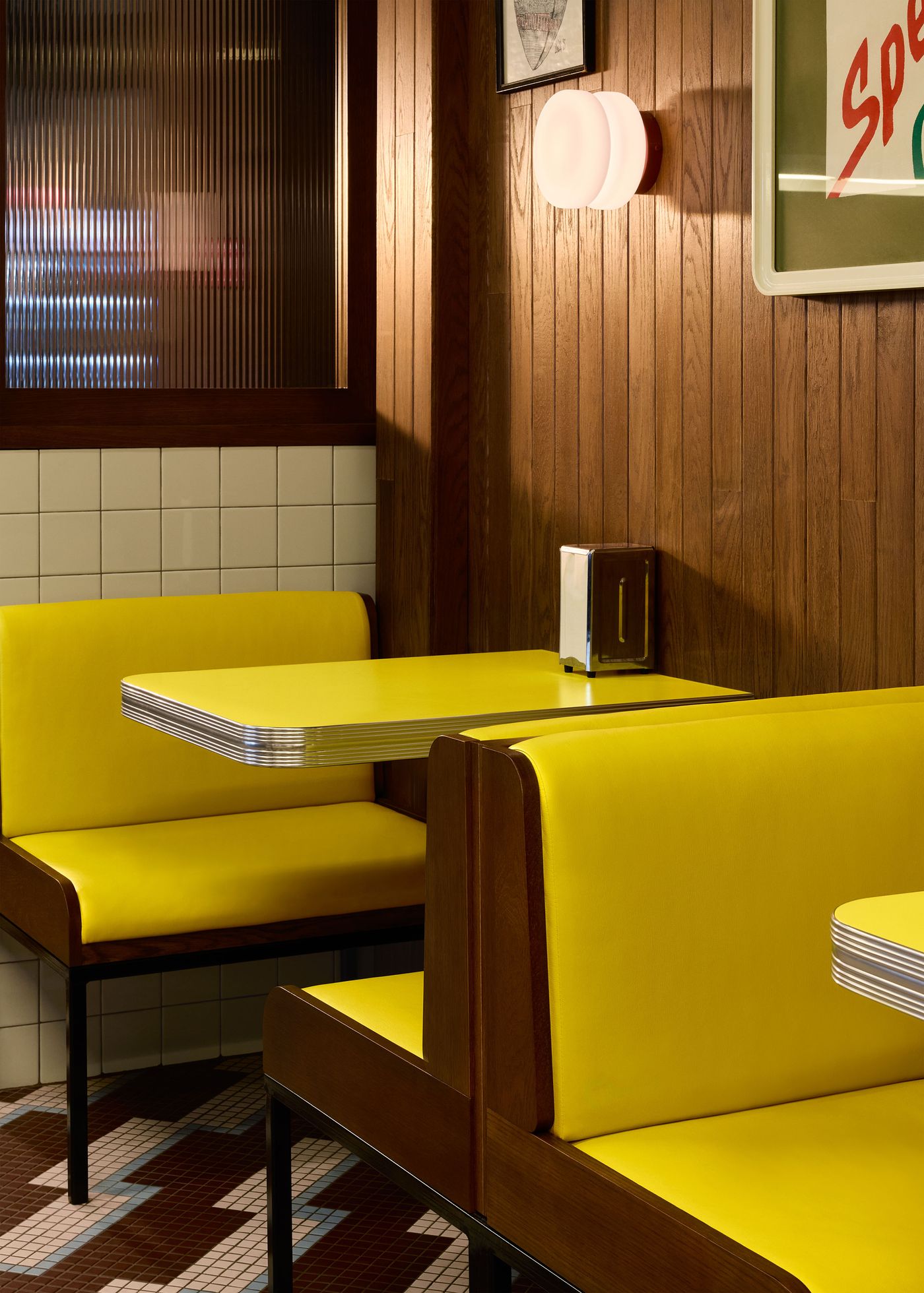 A view of the interior of Renzo Sandwich, featuring bright yellow diner booths and tables. The warm wood-paneled walls, classic checkered tile flooring, and ambient lighting from globe-shaped sconces create a nostalgic and inviting atmosphere. A sign that reads "Spécialité Québécoise" adds a local touch.