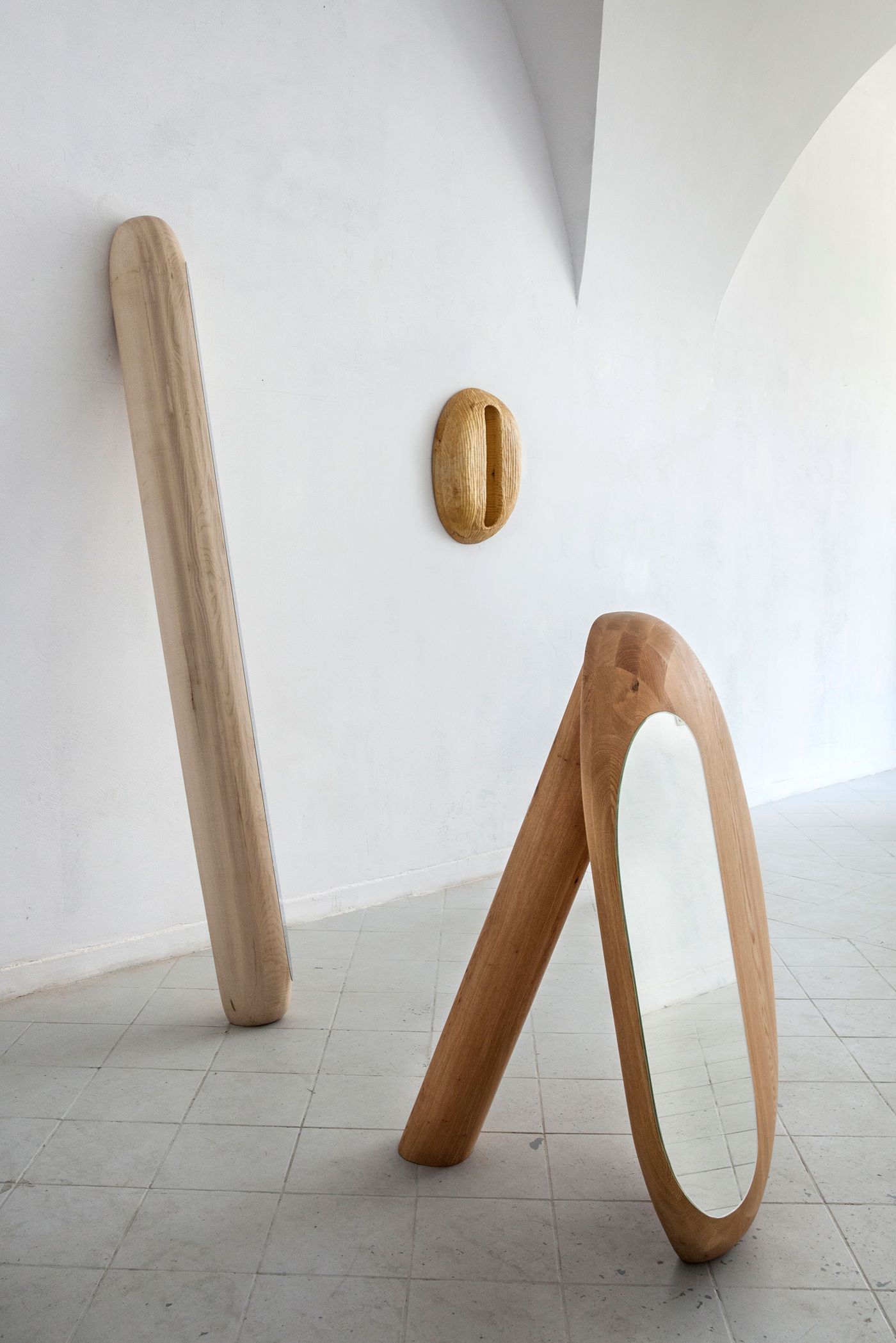 Three abstract wooden sculptures, including a tall mirror and a wall piece, displayed against a minimalist white wall.