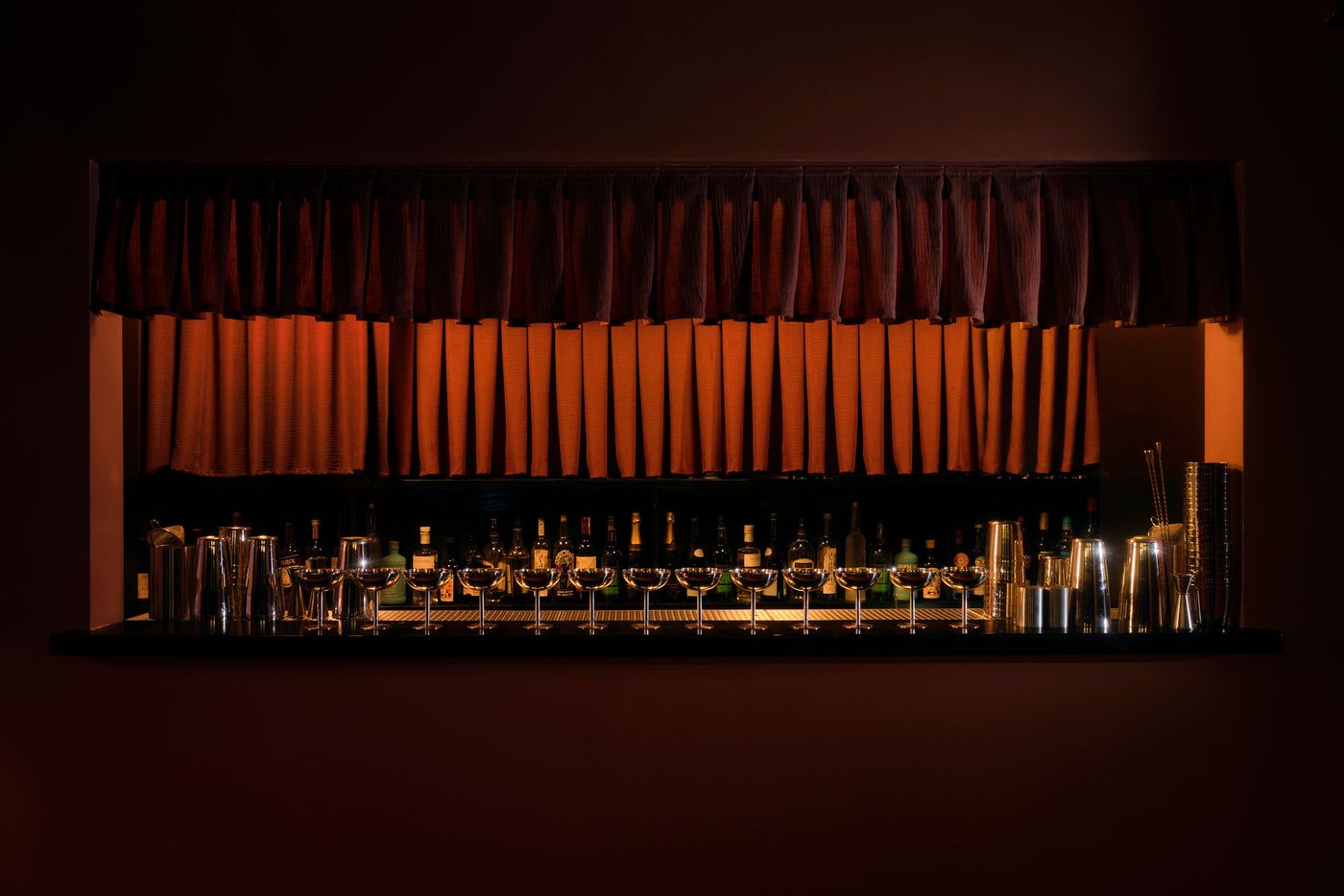A captivating view of the well-stocked bar at The Roses of Elagabalus, framed by rich, pleated red drapes. Below, an array of bottles, elegant stemware, and polished cocktail shakers are meticulously arranged on a dark, reflective surface. The warm, inviting light from behind the drapes enhances the sophisticated and ready-for-service ambiance. A captivating view of the well-stocked bar at The Roses of Elagabalus, framed by rich, pleated red drapes. Below, an array of bottles, elegant stemware, and polished cocktail shakers are meticulously arranged on a dark, reflective surface. The warm, inviting light from behind the drapes enhances the sophisticated and ready-for-service ambiance.