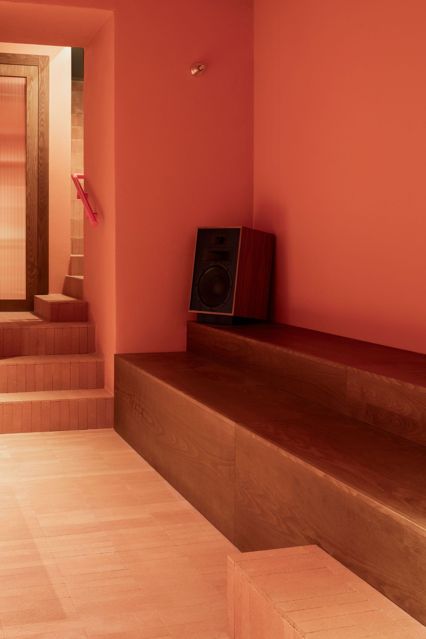 An interior detail of Santo Palato by Naessi Studio, showcasing the rich terracotta hues of the walls and a tiered, dark wood bench seating area, suggesting a relaxed, adaptable space. A black speaker on the bench hints at a vibrant atmosphere, while in the background, a wooden door with fluted glass leads to a staircase, reflecting the restaurant's cohesive design.