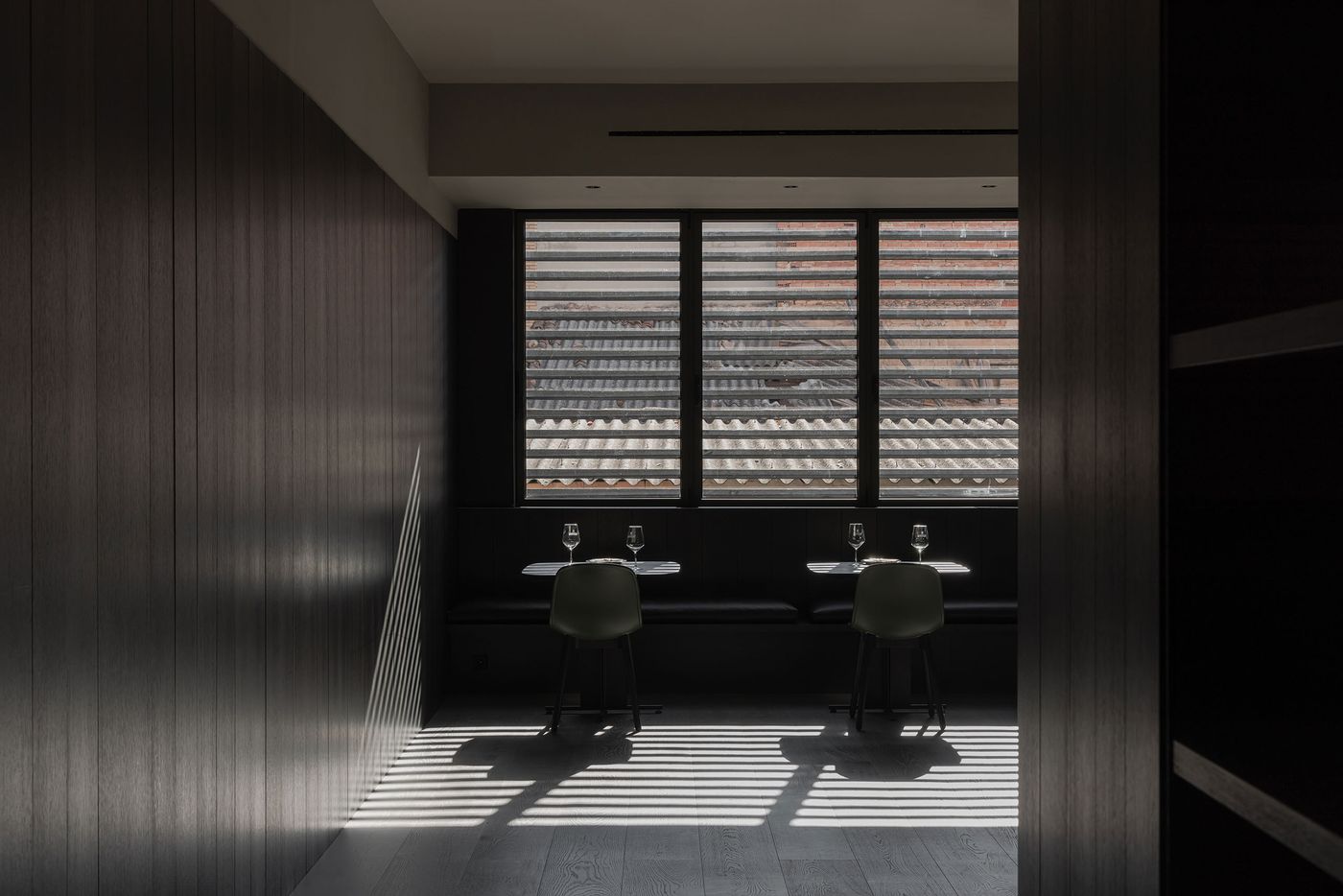 A dimly lit dining space featuring two empty tables set with glasses, showcasing shadow patterns from striped windows.