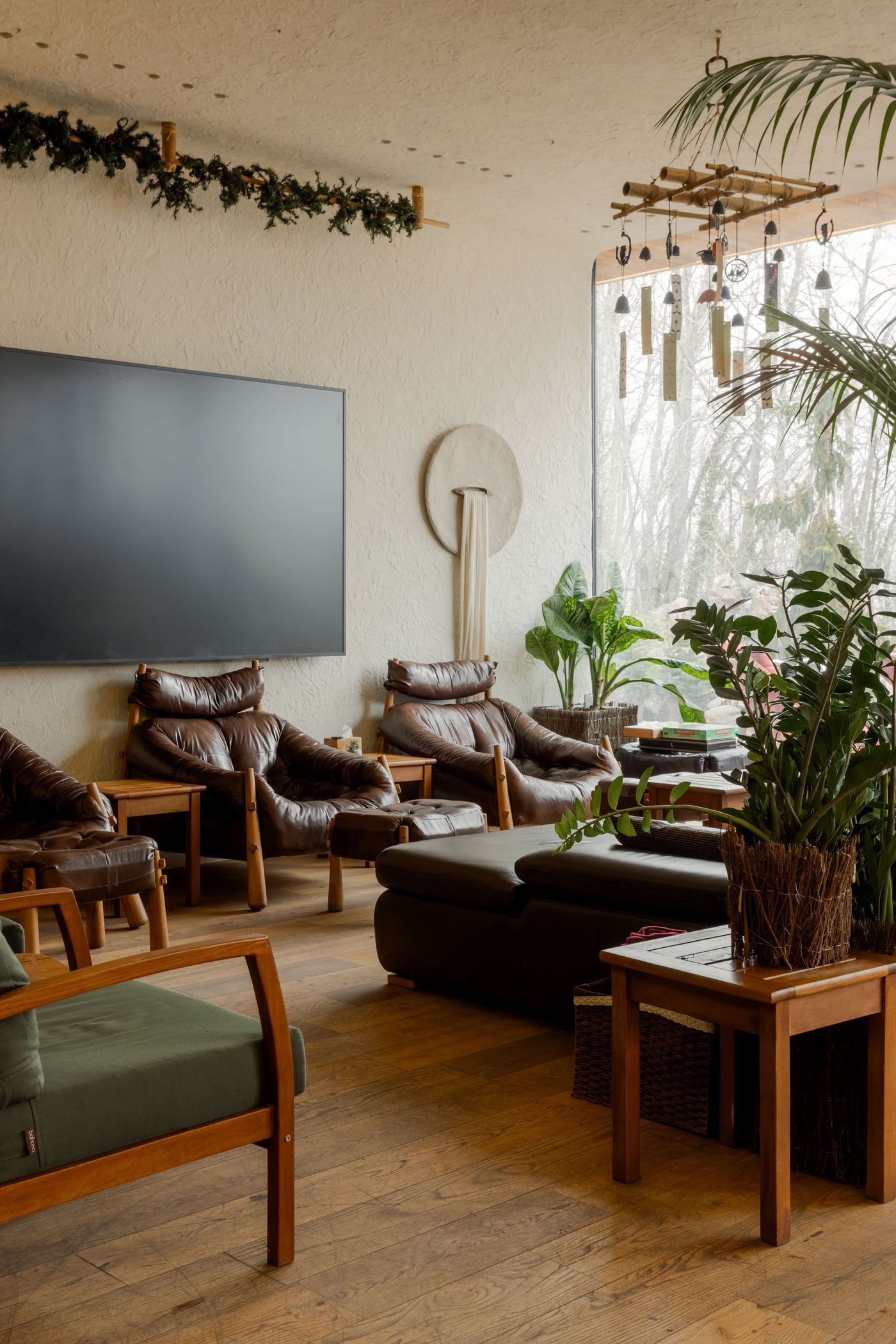 Cozy lounge area with brown leather chairs, wooden furniture, green plants, and a large TV, featuring natural light and rustic decor. Cozy lounge area with brown leather chairs, wooden furniture, green plants, and a large TV, featuring natural light and rustic decor.