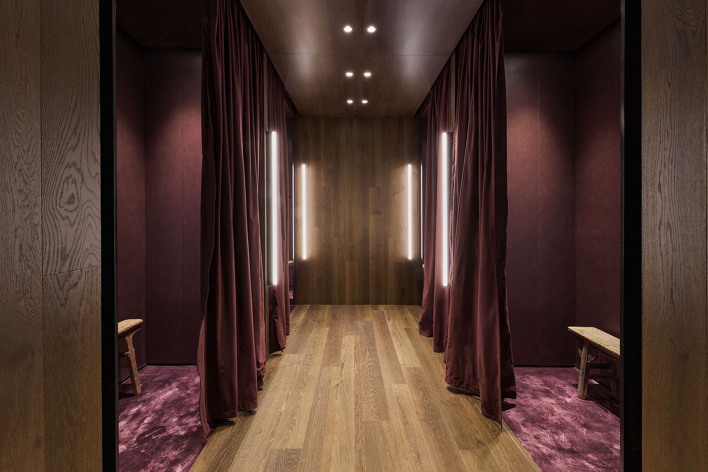 A modern fitting room with burgundy curtains, wooden accents, and warm lighting, featuring a wooden bench and plush purple carpet.