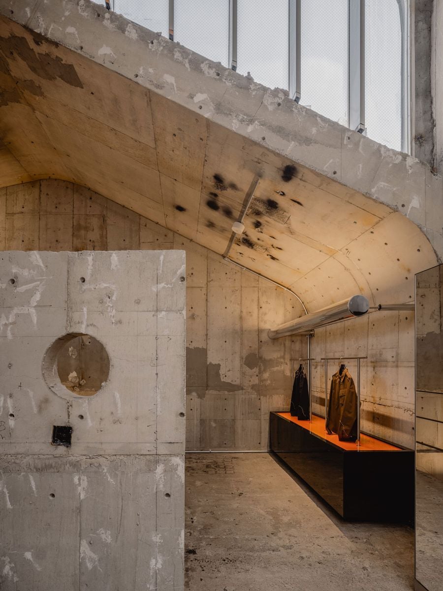 The building’s distinctive concrete ceiling, its stains, patches and construction marks left deliberately exposed. Beneath it, a long black display platform lined in vivid orange supports a sparse arrangement of garments, while a polished chrome tube adds a precise reflective counterpoint to the weathered Brutalist shell.
