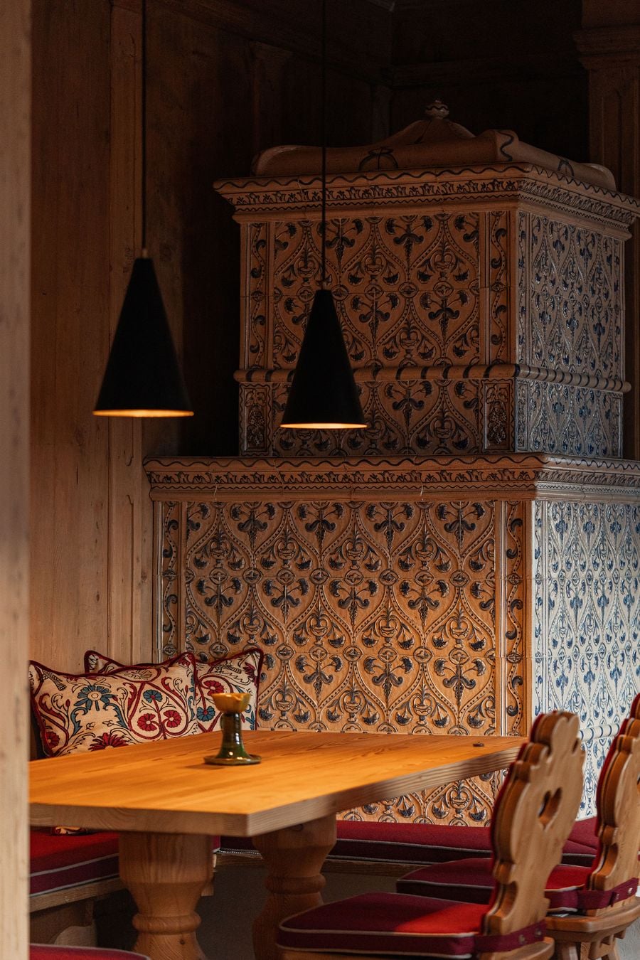 An antique tiled stove dominates the timber-panelled Stube at Castel Badia, its relief tiles patterned with blue ogee and floral motifs. Below, a solid pine table, Tyrolean chairs with carved heart-pierced backs, red bench cushions and embroidered pillows sit beneath two black conical pendants. An antique tiled stove dominates the timber-panelled Stube at Castel Badia, its relief tiles patterned with blue ogee and floral motifs. Below, a solid pine table, Tyrolean chairs with carved heart-pierced backs, red bench cushions and embroidered pillows sit beneath two black conical pendants.
